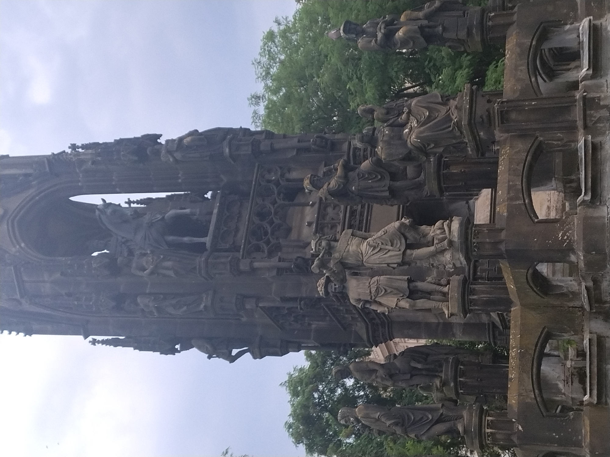 The image shows the National Monument on Vitkov Hill in Prague, Czechia. It is a large, dark stone monument with numerous statues and architectural details. In the foreground, several stone figures are visible, some standing and some seated. One figure on the left appears to be wearing a helmet and carrying a staff. Another figure in the center is holding what looks like a basket of fruit on its head. To the right, a figure is seated with its legs crossed, draped in a robe. The monument itself is tall and imposing, with gothic-inspired architectural elements, including arches and spires. Behind the monument, lush green trees form a dense backdrop against a cloudy, overcast sky. The overall impression is one of historical grandeur and somber monumentality.