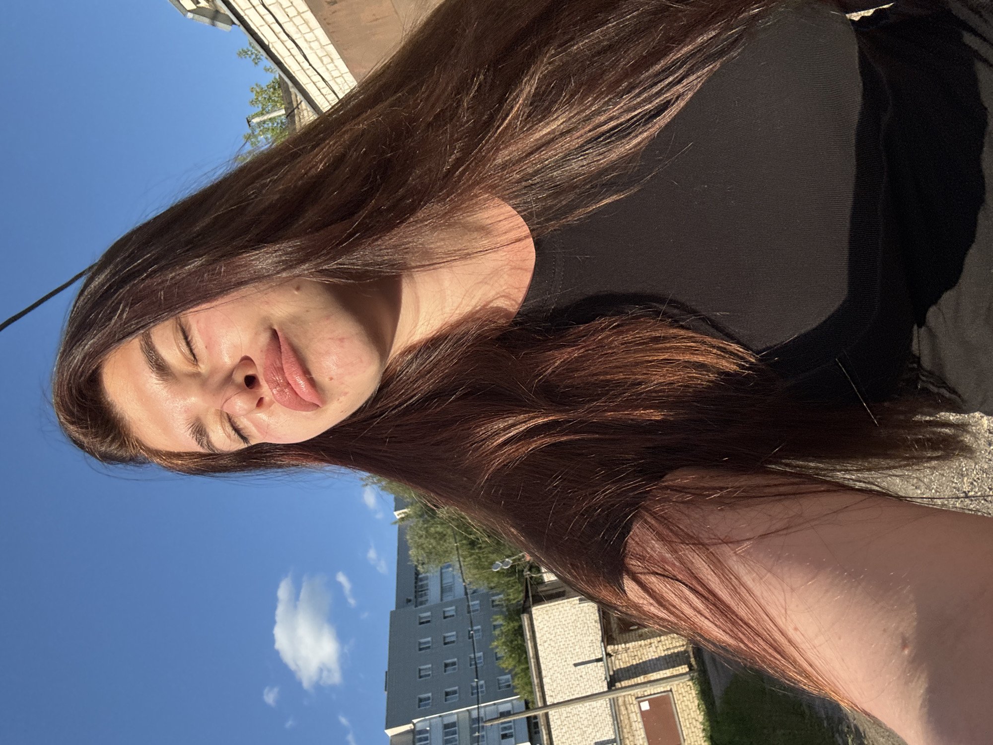 A young woman with long, dark brown hair is taking a selfie outdoors on a sunny day. Her eyes are closed and her lips are pursed. She is wearing a black t-shirt. In the background, there is a blue sky with a few clouds, a multi-story apartment building, and some smaller structures that appear to be garages or sheds. The scene is likely in the city of Klin, Russia, based on the architectural style of the buildings and the overall environment.