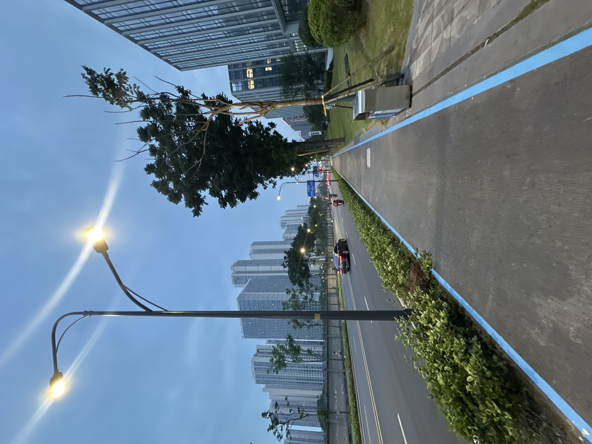 This is a photograph taken in the Chongqing High-tech Zone, China. The scene depicts a street with a pedestrian walkway and a road. The sky is overcast and a pale blue, suggesting it is either early morning or late evening. Two streetlights are visible in the upper left quadrant, emitting a bright yellow light.
The road is asphalt and has two lanes in the direction of travel visible in the frame. A red car is driving on the road, and a motorcycle is also visible further down the street. The pedestrian walkway is to the right of the road, separated by a row of green bushes and a concrete barrier. The walkway is also asphalt, with a bright blue line marking its edge. A silver trash bin is located on the right side of the walkway, next to a grassy area.
On the left side of the road, a series of tall, modern apartment buildings rise into the sky. These buildings are light-colored and appear to be residential. There are trees planted along the road and walkway. On the right side of the walkway, there is a modern office building with a facade of glass and vertical silver accents. The building has windows illuminated with interior lights, indicating occupancy.
The overall impression is one of urban development in a modern high-tech zone, with a blend of residential and commercial buildings, and infrastructure designed for both vehicles and pedestrians.