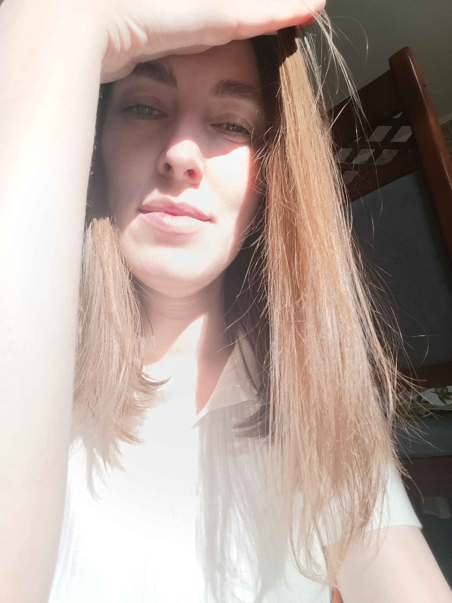 A woman is holding her hand up to shield her eyes from bright sunlight. She is fair-skinned with light brown, shoulder-length hair. She is wearing a white collared shirt. A wooden chair is visible in the background to her right. The photo appears to be taken indoors, with a window as the light source. There is no evidence to indicate the location is in Фоки, Russia.