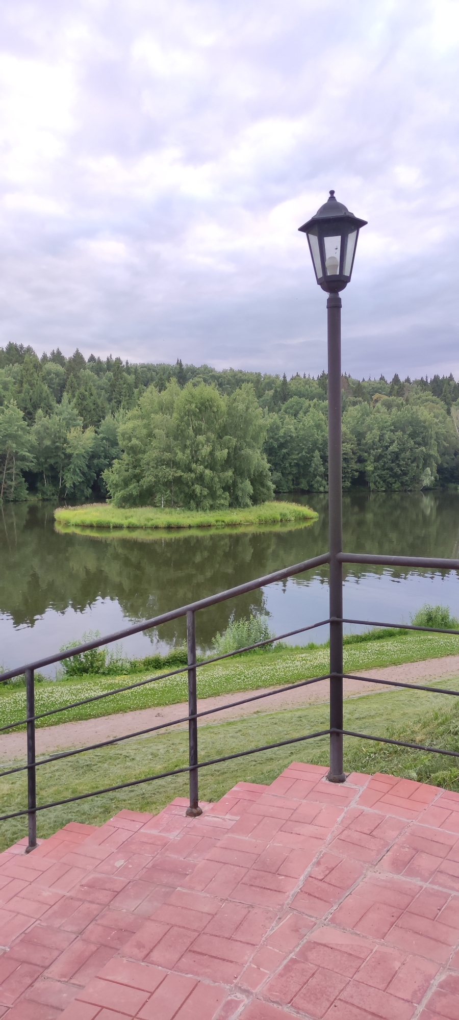 A medium shot captures a serene outdoor scene with a body of water, a small island, a lush forest, and architectural elements. In the foreground, a set of red brick stairs leads to a black metal railing that stretches across the frame, separating the viewer from the water. A black lamppost stands prominently on the right side, its glass panes reflecting the overcast sky. Beyond the railing and the water, a small, verdant island is visible, covered in grass and a cluster of trees. The background is dominated by a dense, green forest that extends across the horizon. The sky is filled with soft, grey clouds, indicating an overcast day. The image appears to have been taken in or around Росинка Кп, Russia, based on the provided context.