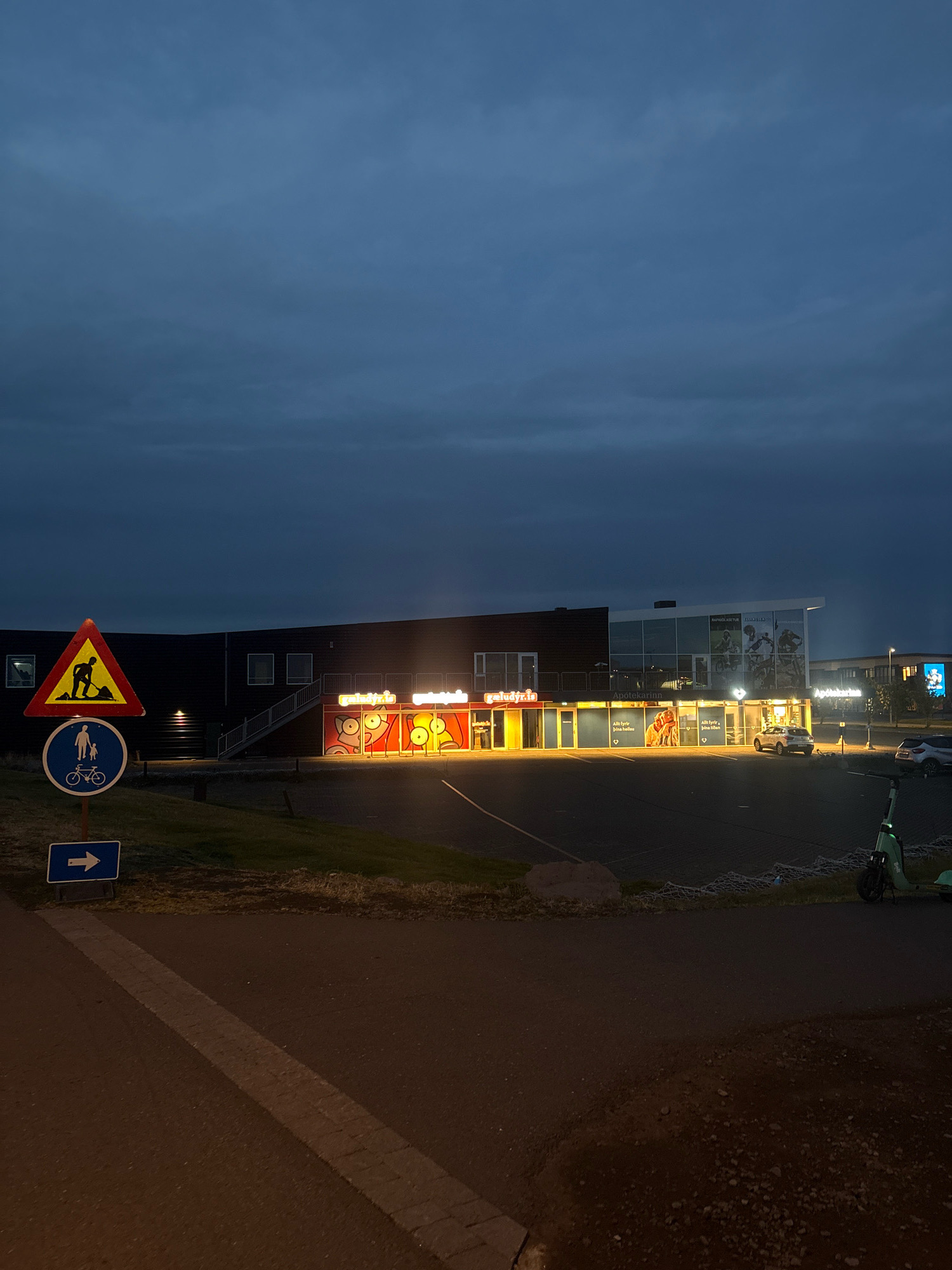 The image captures a dark, cloudy twilight scene in Reykjavik, Iceland, with the prominent facade of a commercial building illuminated by interior lights and signage. To the left, a triangular yellow warning sign with a depiction of a worker digging is mounted above a blue circular sign indicating a shared pedestrian and bicycle path. Below these, a blue rectangular sign points to the right.
The building itself features a dark, modern exterior with a large glass-fronted section on the right. The glass area displays images of cyclists. The left portion of the building has an entrance with illuminated signage that reads "geluðyr.is" and "geluðyr.is" repeated, with red and yellow graphic elements resembling cartoon faces. Further to the right, an area labeled "Apotekarin" is visible, with text below reading "Allt fyrir þína heilsu" (Everything for your health).
Parked cars are visible in the lot in front of the building, and a green electric scooter is parked near the right edge of the frame. The ground is paved, with a striped marking on the asphalt path in the foreground. The overall atmosphere is calm and nocturnal, characteristic of a late evening in a commercial district.