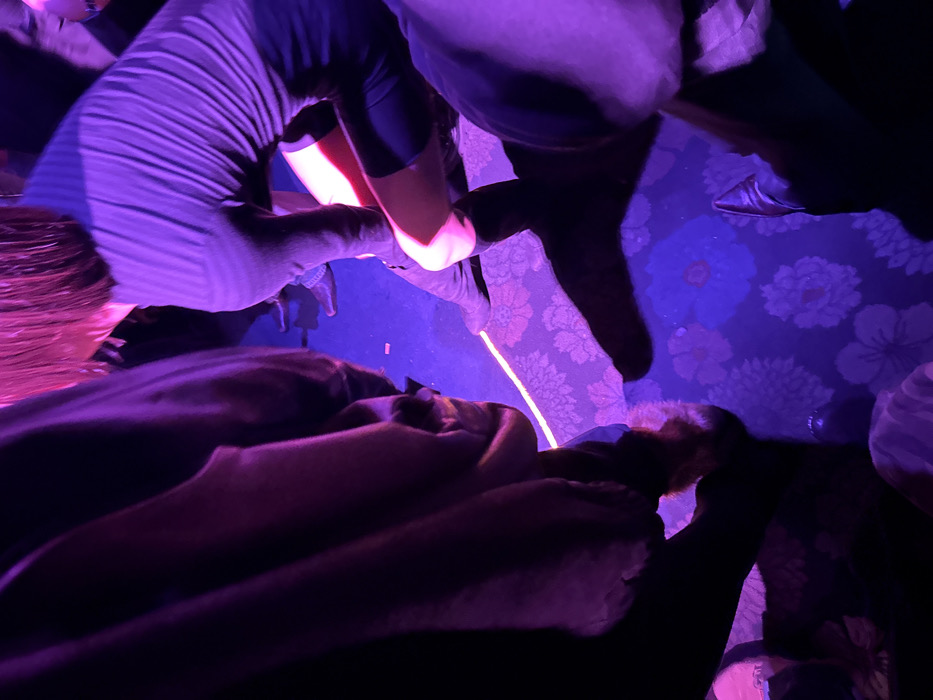 The image depicts a low-angle, close-up view of several individuals within a dimly lit indoor venue, characterized by intense purple and pink artificial lighting. The specific location in Paris, France, cannot be determined from the visual information provided.
In the mid-ground right, a person with short, reddish-brown hair, illuminated by pink light, is observed bending forward and downward. This individual wears a dark, long-sleeved top and dark shorts or a skirt, with their knees and lower thighs prominently highlighted by a bright, reddish-pink light. They appear to be adjusting dark, knee-high boots. To the left, the mid-section and lower body of another person are visible, dressed in dark clothing with a brown fur trim near the bottom edge. In the foreground right, a leg clad in dark trousers and a dark, pointed-toe shoe are present. Additional feet and lower legs are discernible in the background, indicating the presence of a crowd.
The floor is covered with a dark blue or purple carpet featuring a repetitive pattern of lighter purple and white floral designs. A distinct, thin, bright light strip runs in a curved line across the carpet. Small, scattered pieces of debris, possibly confetti, are visible on the carpet. The artificial lighting creates prominent shadows of the individuals on the floor, enhancing the overall atmospheric effect.