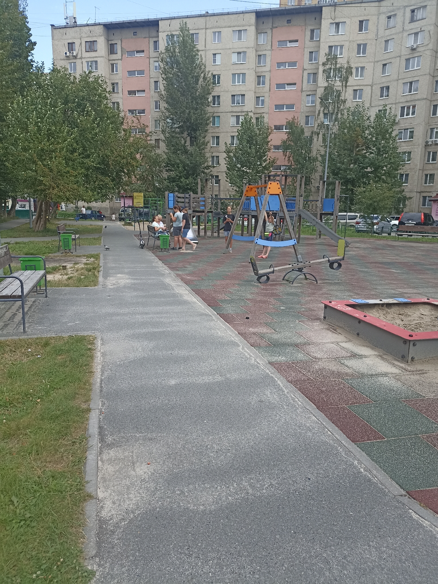 The image depicts a playground area in an urban setting. In the background, a multi-story apartment building, typical of Soviet-era architecture, dominates the skyline. Trees and greenery are interspersed with the building and surround the playground. The playground itself features various equipment including swings, a seesaw, and a sandbox. A few people are present, with some sitting on a bench and others interacting near the play structures. A paved walkway leads through the area, bordered by grass and a playground surface made of rubber tiles. The scene appears to be a public space within a residential district.