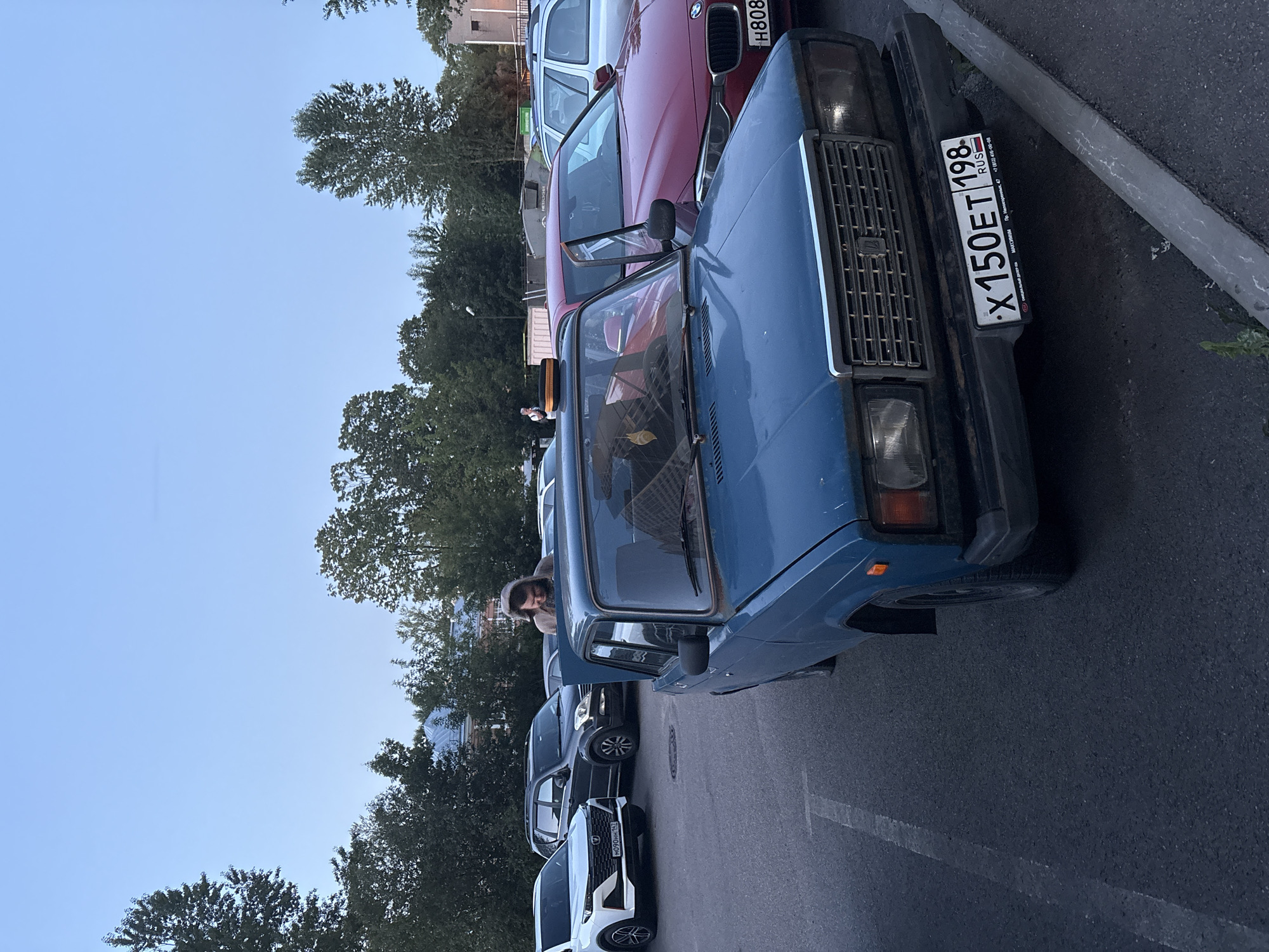 The image depicts a parking lot scene in the city of Imeni Sverdlova, Russia, under a twilight sky. In the foreground, a weathered blue sedan is parked, its hood open. A person wearing a hooded sweatshirt is leaning into the open hood, appearing to be inspecting or working on the engine. Several other vehicles are visible in the background, including a white SUV, a dark SUV, a red sedan, and a silver sedan. Trees and buildings are present in the background, suggesting an urban or suburban environment. The ground is asphalt, with painted parking lines visible. The overall impression is of a casual, everyday moment in a parking area.