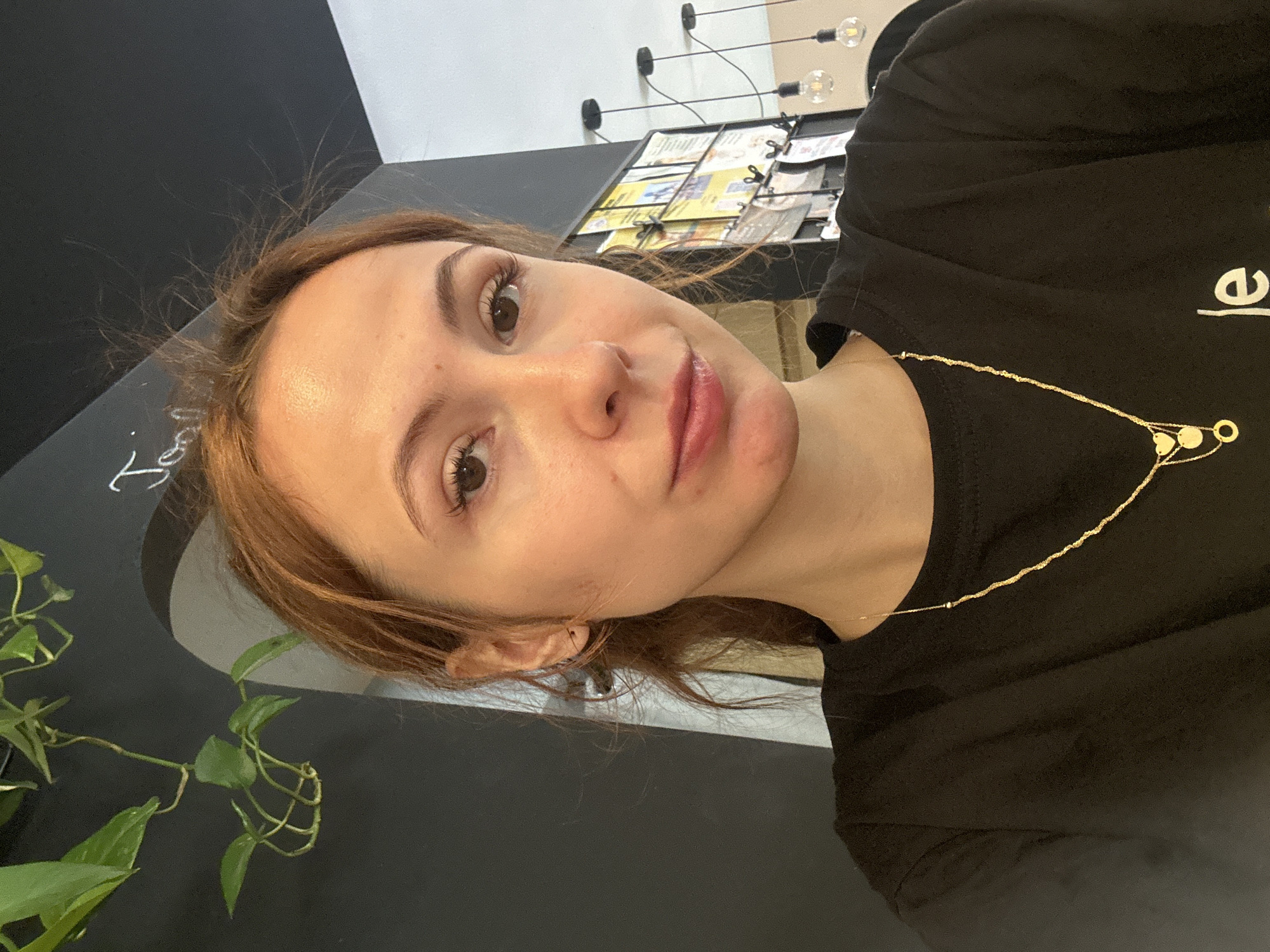 A medium-shot photograph shows a woman with light brown hair wearing a black t-shirt and a gold necklace. The background includes a black wall with a partial inscription in white, a green plant, a lighting fixture with exposed bulbs, and a display of brochures or flyers. The location is likely Novi Sad, Serbia.