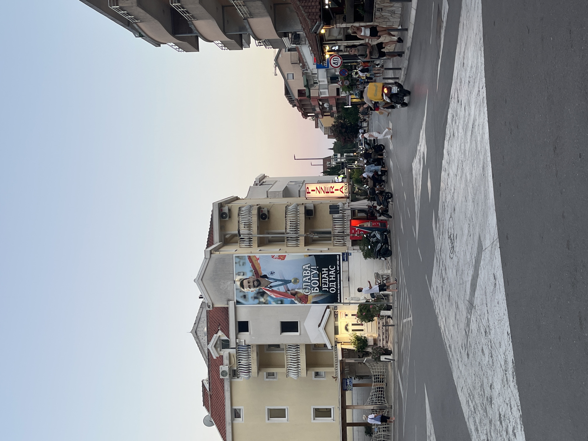 The image shows a street scene in Budva, Montenegro, likely in the evening. The sky is a clear, pale blue, with a hint of pink at the horizon. Buildings line both sides of the street. On the left, a multi-story building with a yellow facade and red tiled roof features a large billboard of an athlete with a medal and a gold necklace, bearing text in Cyrillic. Below the billboard, a pizzeria sign is visible. Several people are walking on the sidewalk and crossing the street. Motorcycles are parked and some are in motion. On the right side of the street, another multi-story building with balconies is visible. Pedestrians are walking on the sidewalk. A scooter with a yellow delivery box is riding away from the viewer. Traffic signs, including a speed limit sign for 40, are present. The street is paved with asphalt and has white pedestrian crossing markings. The overall impression is of a bustling urban environment at dusk.