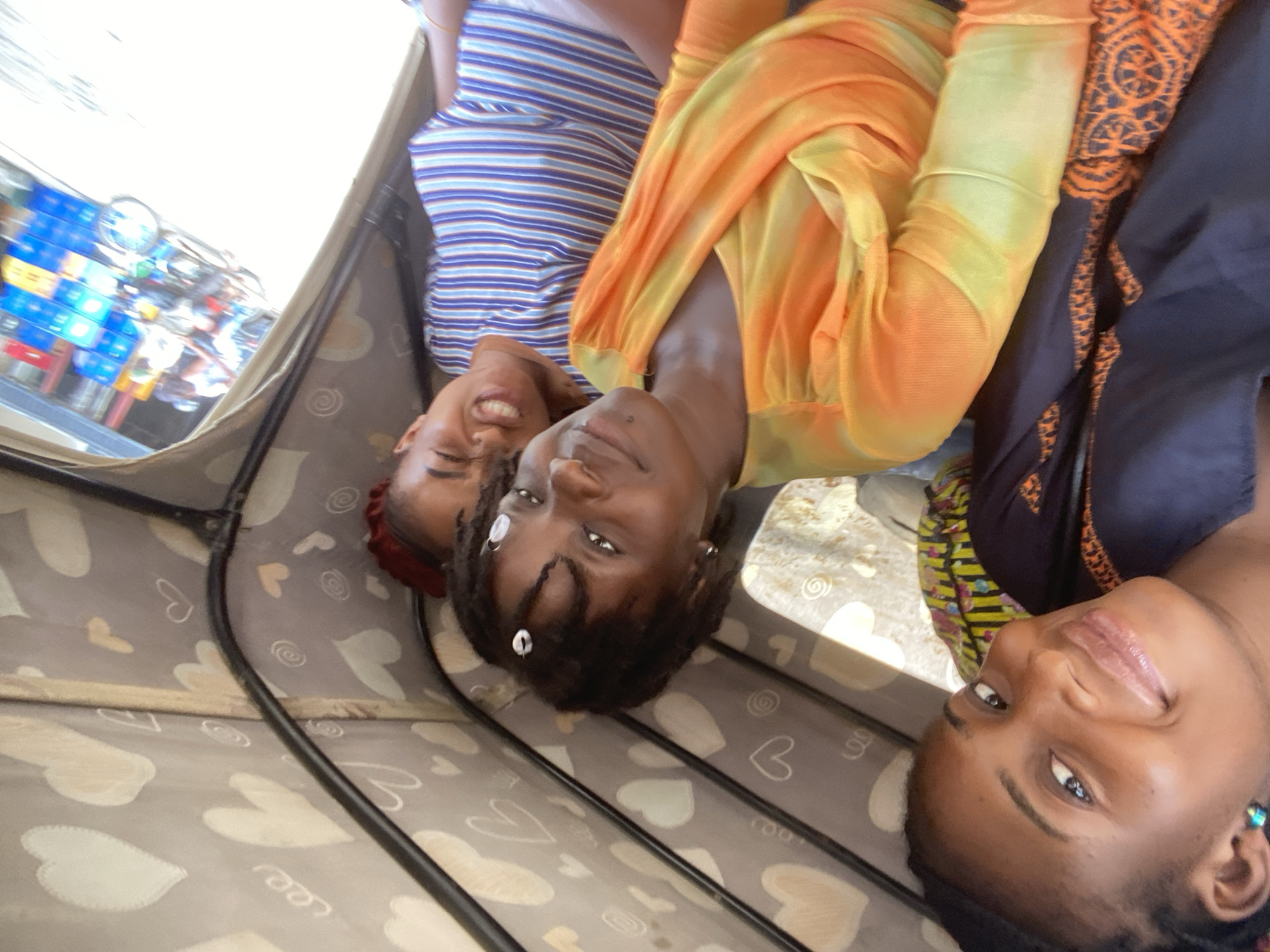 Three young women are captured in a close-up, wide-angle shot from inside a tricycle, commonly known as a keke napep, traveling through a street scene. The interior roof of the tricycle is visible, featuring a grey fabric lining adorned with white and light brown heart shapes and white spiral patterns. Black metal support bars of the vehicle frame traverse the upper section of the image.
The woman on the far left, partially cropped, has dark skin, braided hair, and a slight smile, looking directly at the camera. She wears a dark blue garment with an orange embroidered or patterned design. A small, vibrant turquoise earring is visible in her left ear.
The central woman has dark skin, short, dark dreadlocks or natural twists, some of which are decorated with white cowrie shells or beads. She has a gentle smile and is looking directly at the camera. She wears a long-sleeved top with a draped neckline, featuring a vibrant tie-dye pattern in shades of yellow and orange. A small silver hoop earring is visible in her left ear.
The woman on the right, also with dark skin, exhibits an open-mouthed, expressive smile or grimace. Her head is partially covered by a red headwrap or cap. She wears a blue and white horizontally striped t-shirt. Her gaze is directed slightly past the camera.
Through the open side of the tricycle on the right, a blurry but distinct street scene is visible under bright daylight. Elements include a person riding a motorcycle (okada), another individual and a vehicle, and prominent stacks of blue and some red plastic crates, suggesting a commercial or market area. The overall scene depicts a common street and transit environment typical of urban settings in Abuja, Nigeria, characterized by local transportation and roadside commerce.