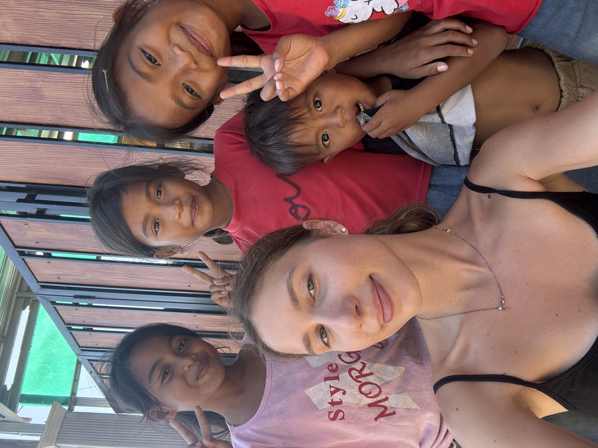 A group of five people, including one adult woman and four children, pose for a selfie outdoors. The woman, with light skin and brown hair pulled back, is in the foreground. She wears a black tank top and a delicate necklace. Behind her, three young girls and one young boy are visible. The girls, with dark hair and eyes, wear t-shirts in pink and red. The boy, also with dark hair and eyes, wears a grey and white shirt and appears to be holding something to his mouth. The background consists of a wooden fence and a glimpse of a green roof structure. The setting is likely Kertasari, Indonesia, given the context provided, suggesting a casual, outdoor gathering. The overall mood is cheerful and friendly, with several individuals flashing peace signs.