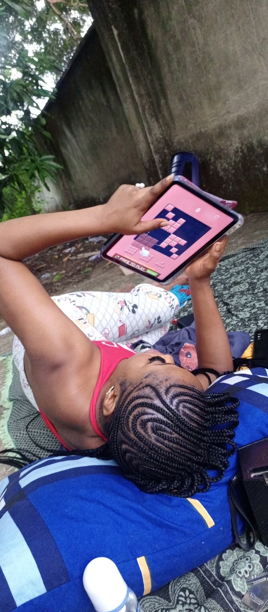 A person is lying outdoors in Keffi, Nigeria. They are lying on top of a blue striped pillow, on top of a dark green patterned blanket, wearing a red tank top and light-colored Mickey Mouse-patterned pants. They are holding a tablet with a pink border and a puzzle game displayed on the screen, using their finger to interact with the screen. The background consists of a weathered gray concrete wall and some foliage.