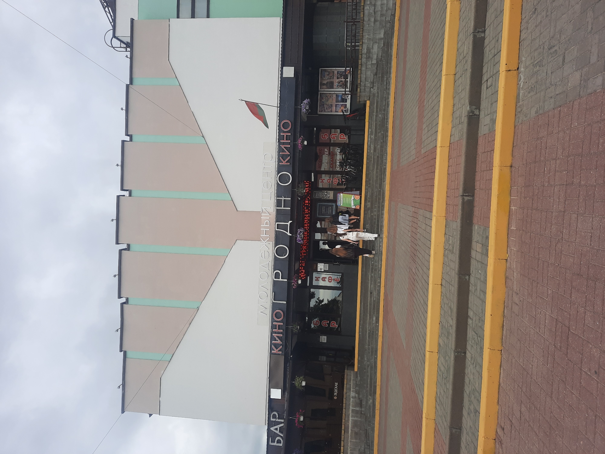 The image depicts the entrance of the "Molodezhnyy Tsentr" (Youth Center) in Hrodna, Belarus. A large, multi-story building dominates the frame, with a facade featuring distinct colored panels and a prominent illuminated sign spelling out "МОЛОДЕЖНЫЙ ЦЕНТР" and "КИНО ГРОДНО КИНО." A Belarusian flag flies from a pole above the entrance.
The ground level of the building houses a dark-colored entrance area with multiple signs, including "БАР" (Bar), "КАФЕ" (Cafe), and "КИНО" (Cinema). Several people are seen entering and exiting the building. Two women, one in a white top and pants and the other in a white dress and pants, are walking up a wide set of stairs leading to the entrance. Other individuals are partially visible inside the entrance. Bicycles are parked near the entrance.
The foreground shows a paved walkway with distinct red and grey brick patterns, bordered by yellow curbs. A series of steps, also with yellow borders, leads up to the building's entrance. The sky above is overcast with a neutral grey tone. The overall scene suggests a public gathering place or entertainment venue on an ordinary day.