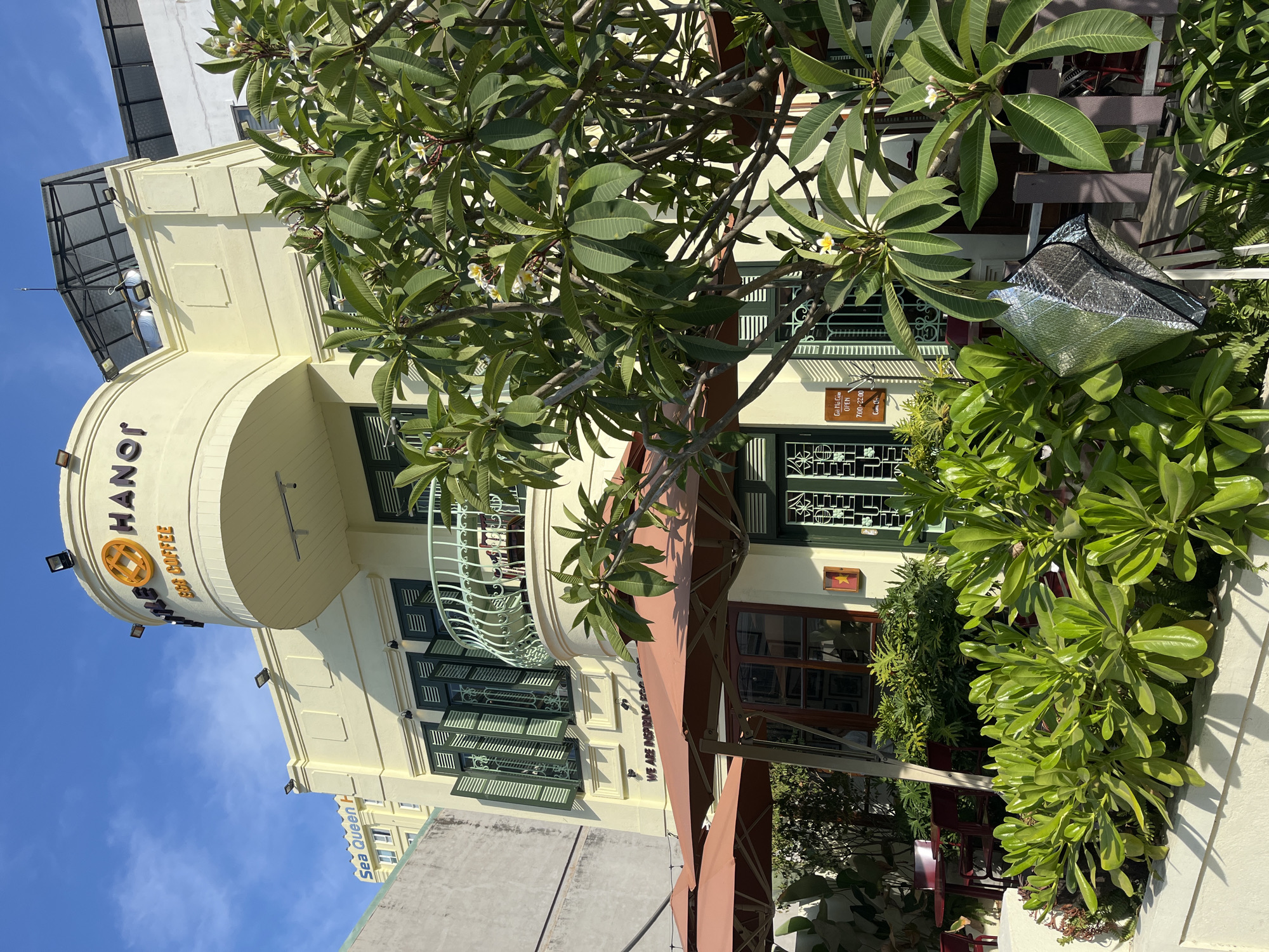 The image presents a sunny daytime view of a charming, multi-story building in Đà Nẵng, Vietnam, likely a café or restaurant. The building's architecture features a light yellow-cream facade with traditional dark green shuttered windows and a prominent curved corner. A decorative balcony with white railings is visible on the upper floor, where a person is partially seen looking outwards, suggesting a relaxed atmosphere.
Lush green foliage, including trees with white and yellow frangipani flowers and various potted plants, frames much of the scene, creating a vibrant and natural ambiance. Bright sunlight casts distinct shadows, indicating clear weather. An awning extends over an outdoor seating area on the ground floor, where tables and chairs are partially visible.
Visible text on the curved building section includes "HANOI COFFEE" with its distinctive logo. A small wooden sign near the entrance reads "Giờ Mở Cửa" (Opening Hours), "OPEN", and "7:00 - 22:00". On an adjacent building, the text "SEA QUEEN" is visible. Part of a slogan, "WE ARE INSPIRING BO", is seen on the awning. A metallic insulated bag hangs among the plants, and a small Vietnamese flag emblem is displayed on the wall. The scene captures a peaceful and inviting urban setting, characteristic of a pleasant café experience.