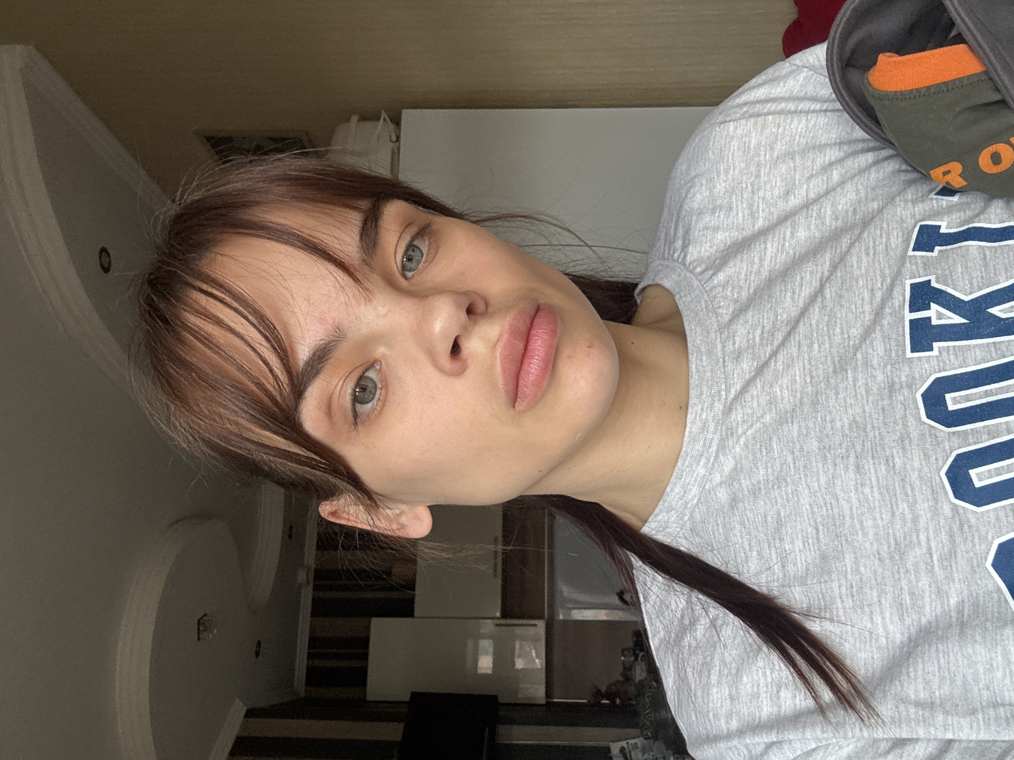 A young woman with fair skin, brown hair, and green eyes is facing the camera. She is wearing a light gray t-shirt with large blue lettering on it. A hooded sweatshirt with orange accents is visible on her right shoulder. The background includes a white refrigerator, an off-white ceiling with decorative molding, and a glimpse of a striped rug or flooring. There is no definitive information provided in the image to place the location specifically in Rostov-on-Don, Russia.