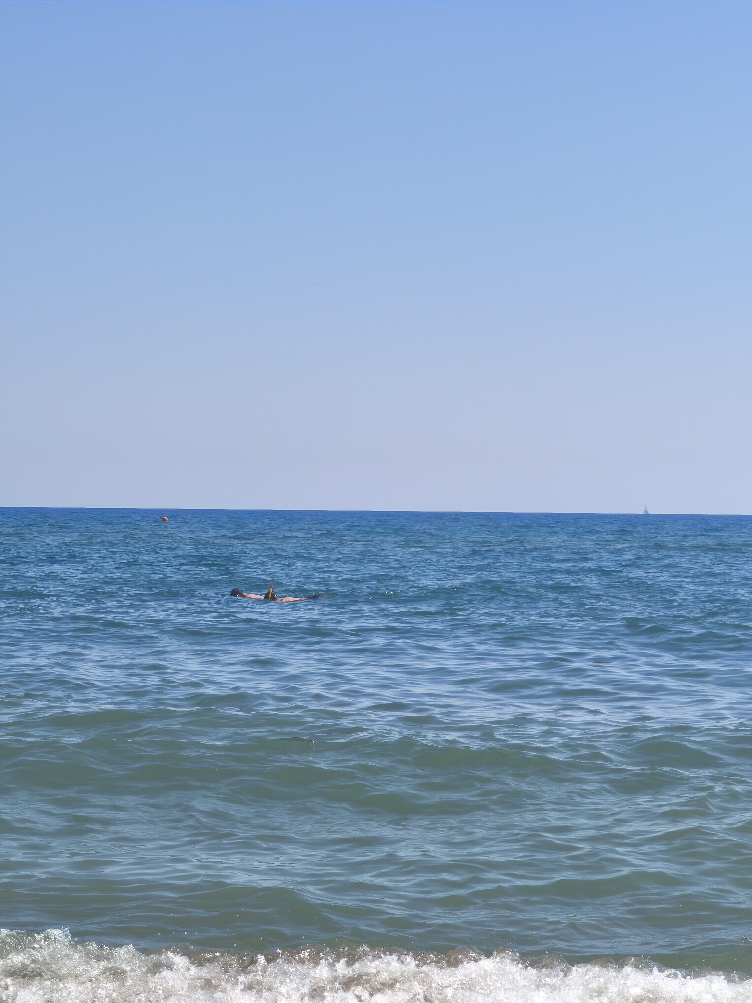 The image depicts a tranquil scene of the sea at Castiglione della Pescaia, Italy, under a clear blue sky. In the middle ground, a person is seen floating on their back, wearing a snorkel mask and fins, suggesting they are engaged in snorkeling or leisurely floating. The water is a deep blue, with gentle waves rippling across its surface. A small orange buoy is visible in the distance to the left, and a distant sailboat can be spotted on the horizon to the right. The foreground features the edge of a breaking wave with white foam washing onto what appears to be a sandy shore, though the shore itself is not clearly visible in this frame. The overall impression is one of a calm and sunny day at the beach.