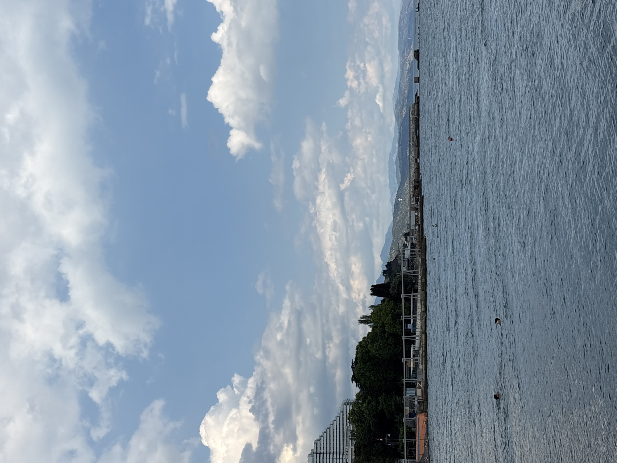 The image depicts a waterfront scene in Alushta, Ukraine, under a sky filled with large cumulus clouds. In the foreground, the dark blue water of the sea is disturbed by ripples. Two individuals are visible in the water, with their heads above the surface, appearing to be swimming or treading water. A red buoy floats further out in the sea.
Along the coastline, a pier or promenade stretches horizontally. Behind the pier, lush green trees form a dense line. A prominent, pyramid-shaped white building, possibly a hotel or resort, stands to the left, its angular structure contrasting with the natural landscape. Further in the background, a range of hills or mountains form a hazy silhouette against the sky. The scene suggests a recreational area with a combination of natural beauty and modern architecture.