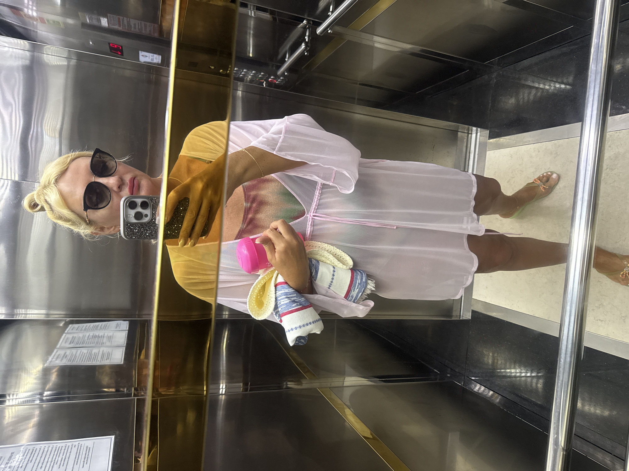 A woman with blonde hair pulled into a bun, wearing large dark sunglasses, a sheer pink cover-up over a tie-dye swimsuit, and flip-flops, stands in a mirrored elevator. She is holding a pink water bottle and a rolled-up towel or sarong. The elevator has stainless steel walls and a gold-colored handrail. The elevator number displayed on a panel is "19". The woman is taking a selfie with her phone. The scene appears to be in a hotel or apartment building. The location is Alanya, Turkey.