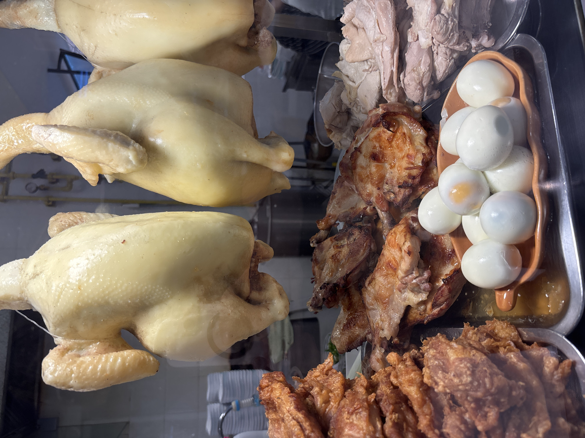 The image depicts a food display in บ้านบางรักษ์, Thailand. Three whole, pale yellow cooked chickens are displayed on the upper left. To their right is a metal tray containing sliced, pale meat, possibly boiled chicken. Adjacent to this is another metal tray with grilled chicken pieces. A brown bowl containing white boiled eggs sits on the same tray. Beneath the grilled chicken, a tray holds deep-fried pieces of meat. The display is behind glass, presumably in a restaurant or food stall.