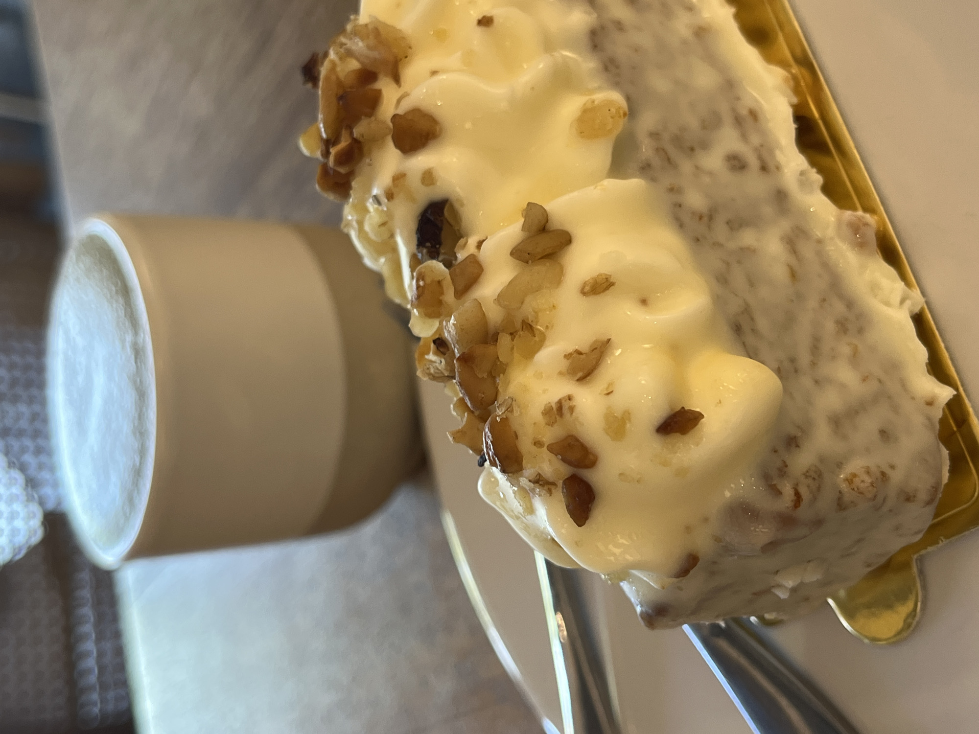 A close-up shot depicts a piece of cake and a mug, potentially in Kuta Selatan, Indonesia. The cake is rectangular and topped with a thick layer of white frosting and chopped nuts. It sits on a golden serving tray or plate and is being lifted or cut with a fork. A beige mug is visible to the side of the cake, partially out of focus. The setting appears to be a casual environment, perhaps a cafe or restaurant.