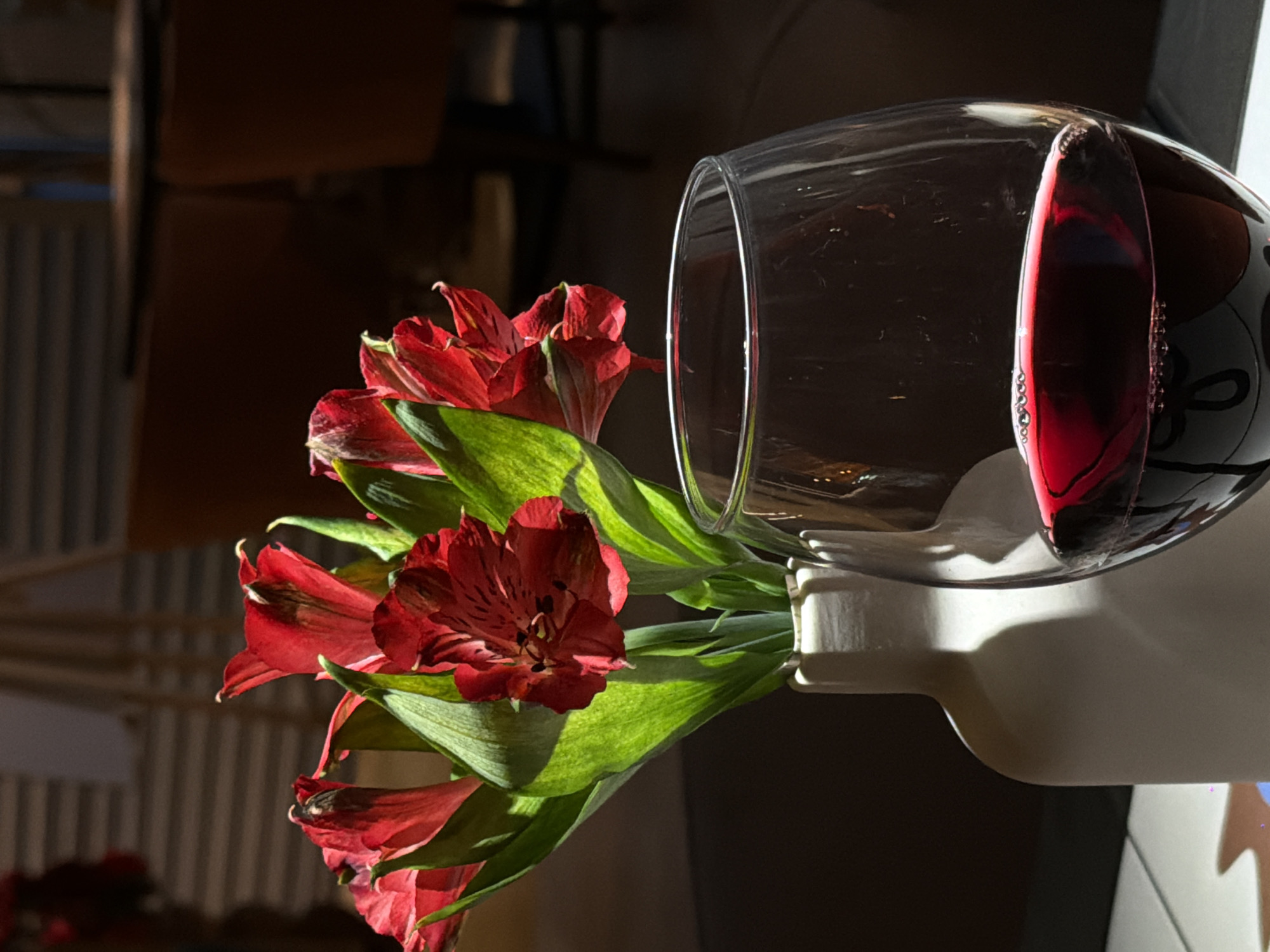 In a dimly lit room, a white vase holds a bouquet of vibrant red lilies with green leaves. To the right of the vase, a stemmed wine glass is filled with deep red wine, with a small amount of foam on the surface. The background is blurred, showing hints of furniture and a striped wall. The overall scene suggests an intimate setting, possibly a restaurant or a private dining area. It is not possible to determine the location within the city of Perm, Russia, from the provided image.