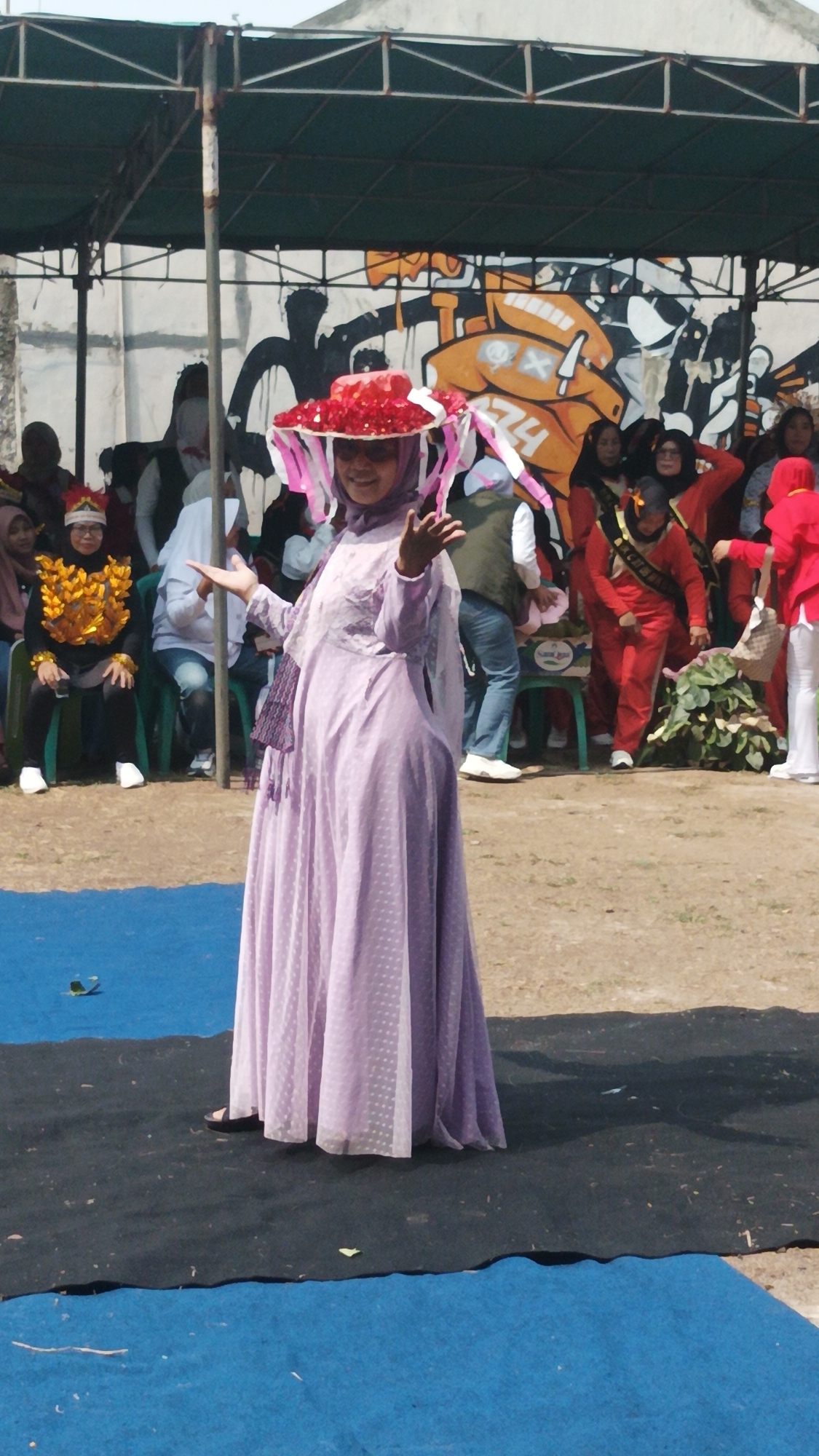 In Munjul, Indonesia, a woman in a purple dress and a wide-brimmed red hat adorned with pink ribbons stands on a black mat, her arms extended. She appears to be performing on a stage or platform, with a crowd of spectators seated in the background. The backdrop features a mural with graffiti art. The event is likely a cultural celebration or festival, possibly a parade or competition, given the decorative attire and the presence of an audience.