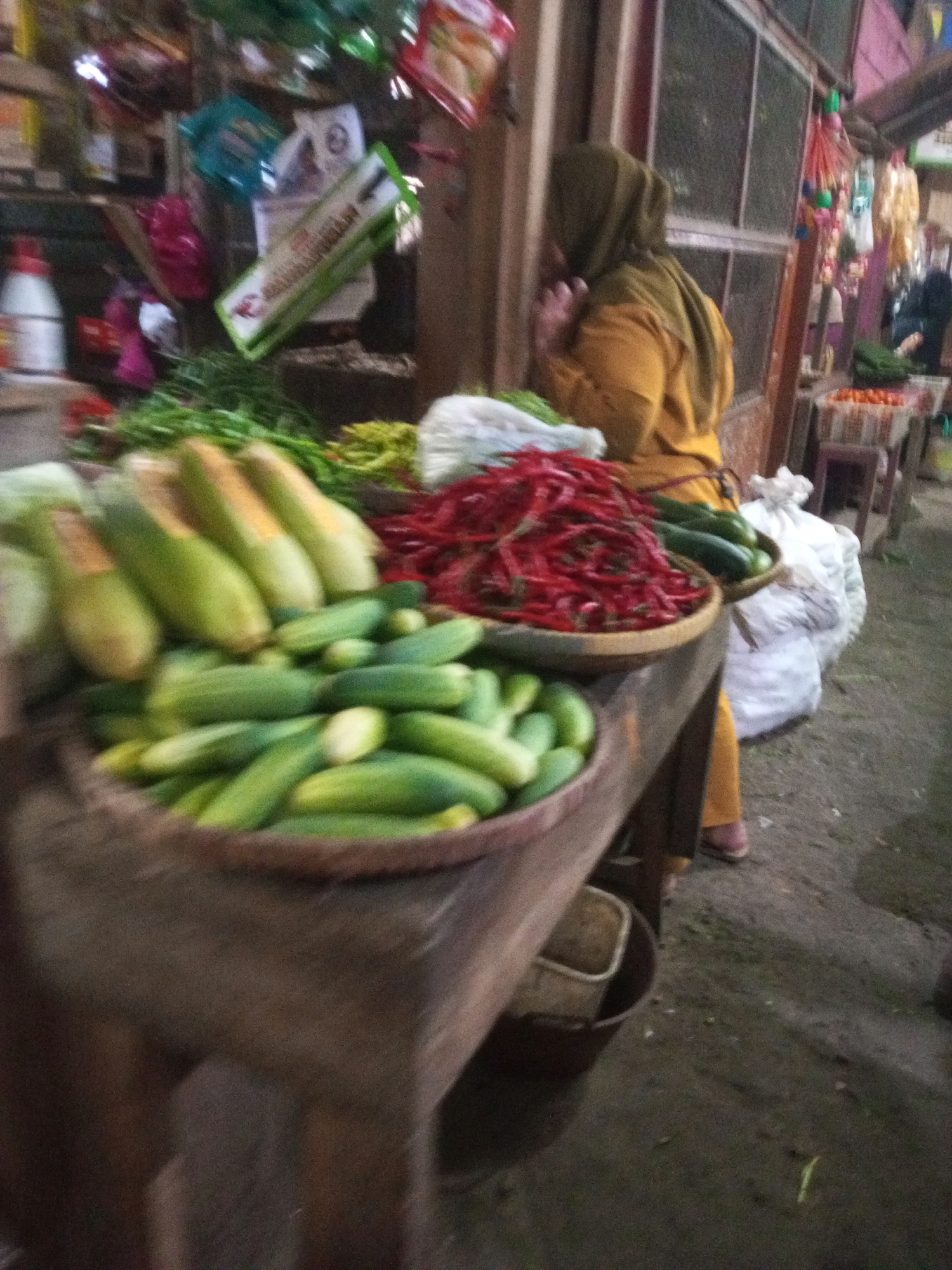 This image captures a scene at a street market in Kalibeber, Indonesia. In the foreground, a wooden table displays a variety of fresh produce. A large bowl or basket is filled with numerous cucumbers, some of which are elongated and pale green, while others are shorter and more vibrant green. Behind the cucumbers, a pile of bright red chilies is visible in a woven bowl. To the left of the chilies, there are bundles of green leafy vegetables, possibly herbs or other produce, with some yellow items also present.
In the background, a woman wearing a dark green hijab and a mustard-yellow garment is seen from the side, facing away from the camera and towards the market stalls. She appears to be interacting with the market or customers. Behind her, market stalls are set up with various goods, including what appear to be packaged items on shelves and some hanging decorations. White sacks are piled on the ground to the right of the woman, and the ground itself is dirt or gravel. The overall lighting suggests it is daytime, possibly late afternoon or early evening given the shadows. The market environment appears lively and busy.