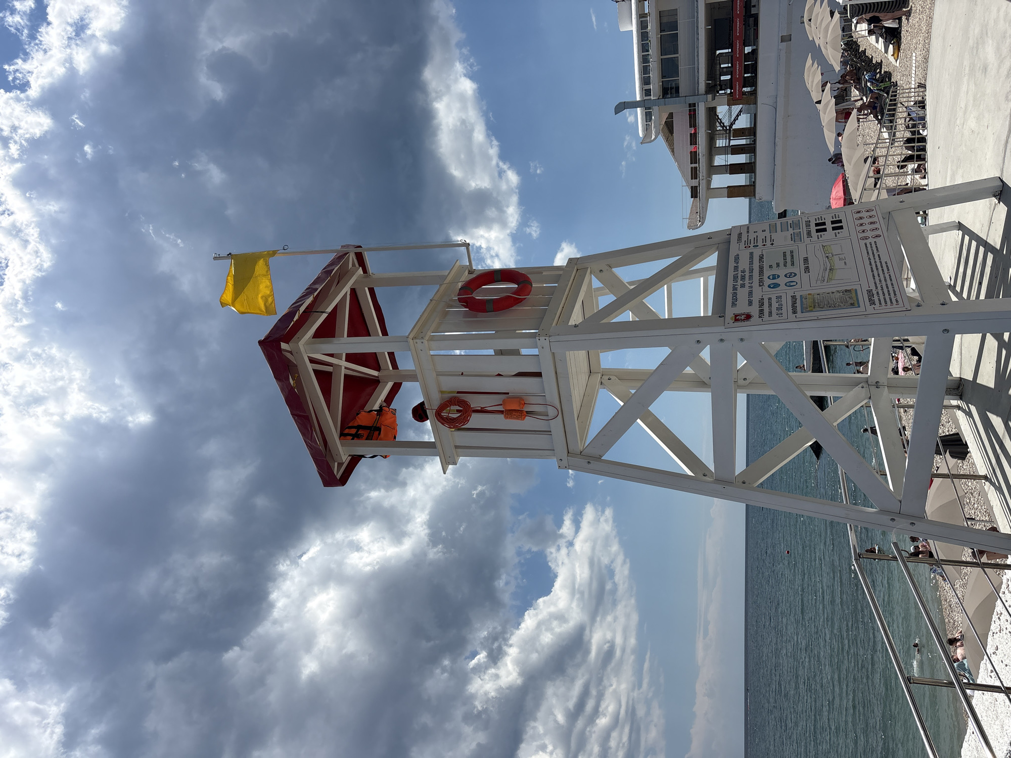 A lifeguard tower with a red roof stands on a pebble beach in Alushta, Ukraine. The tower is white with a ladder and has a yellow flag flying from a pole. Two red life preservers and an orange life jacket are visible on the tower. A sign with information and rules in Russian is attached to the tower. The sea is to the left, with a few people swimming. To the right, beach umbrellas and lounge chairs are set up, and people are sunbathing. A white building, possibly a hotel or restaurant, is behind the beach umbrellas. The sky is partly cloudy, with a mix of dark and light clouds.