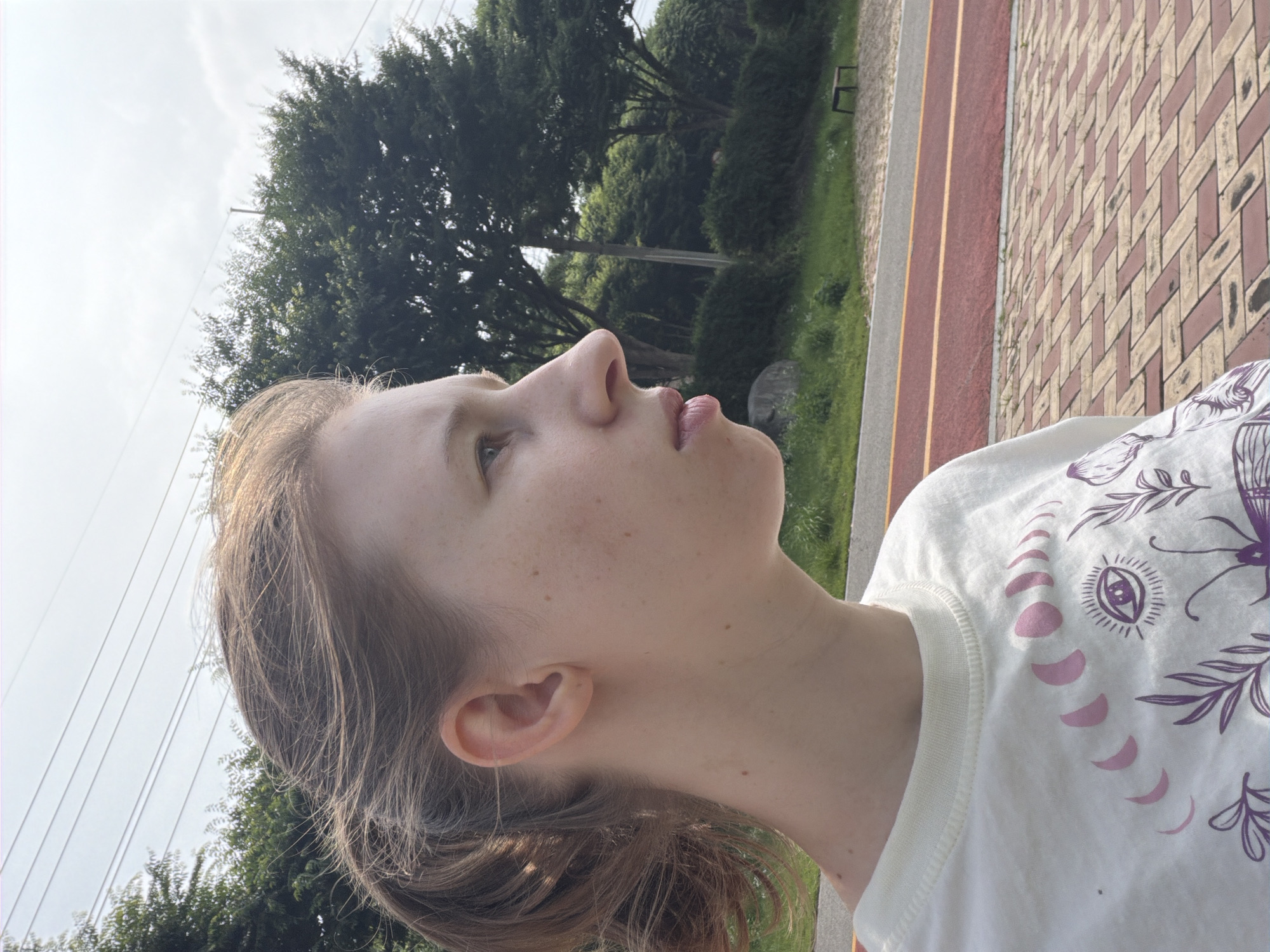 The image shows a young woman in profile, facing right, outdoors. She has light brown hair pulled back and visible freckles on her face. She is wearing a white t-shirt with a graphic print of celestial phases and botanical elements. The background features a paved path with red and yellow markings, green grass, trees, and a cloudy sky. In the far background, a small, dark bench is visible. The scene appears to be a park or recreational area. The location is identified as Dongducheon-si, South Korea.