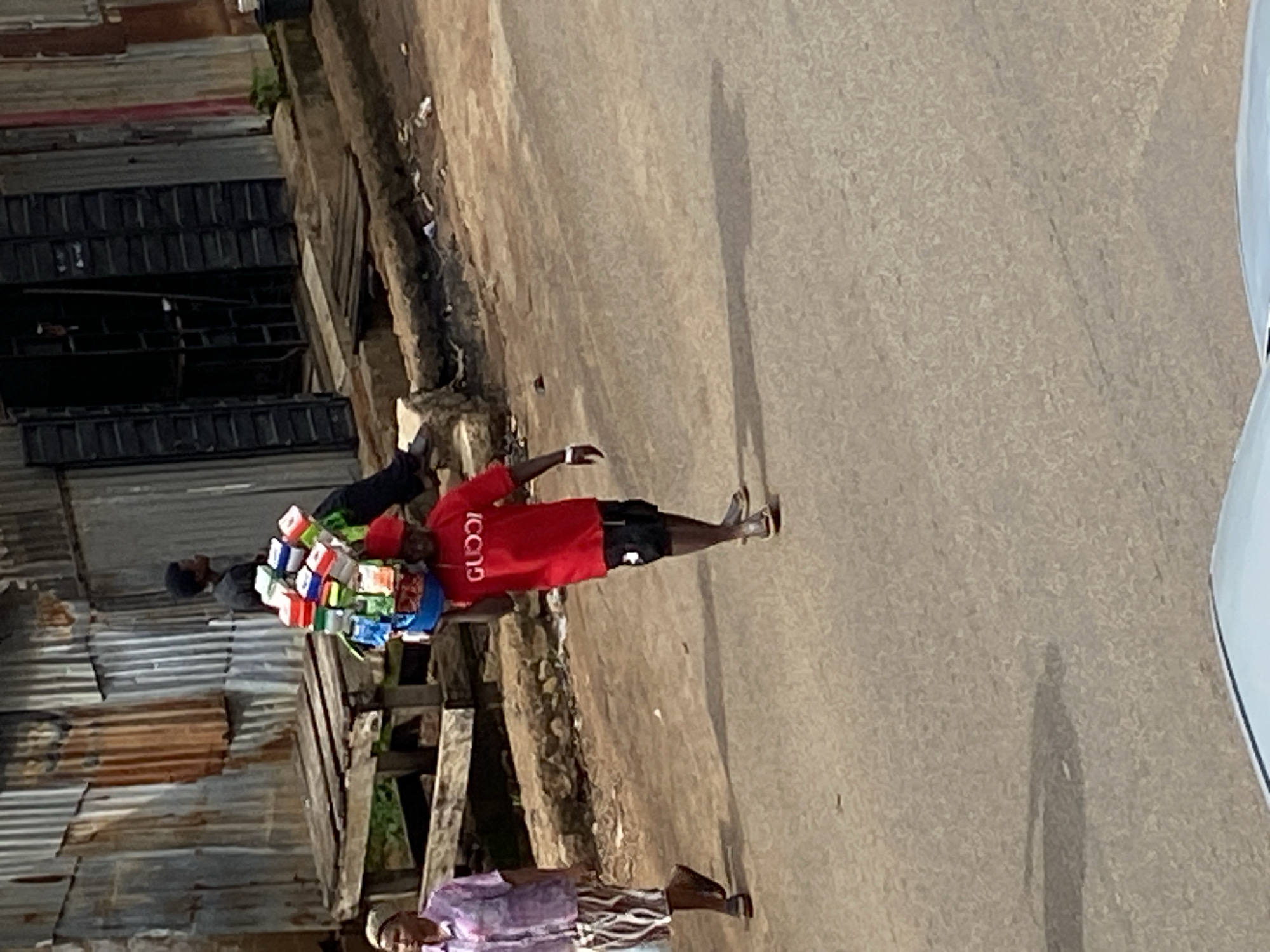 A vendor, identified by the "GUCCI" logo on his red t-shirt, carries a large stack of merchandise, likely soap or detergent bars, balanced on his head. He is walking on a paved street in Bukuru, Nigeria. He wears black shorts and sandals. To his left, a woman in a purple patterned blouse and striped skirt walks in the same direction. Behind them, a corrugated metal structure serves as a backdrop, with a wooden platform and some greenery visible. Another person, wearing a black shirt, is partially visible near the metal structure. The environment suggests an urban setting with signs of commercial activity.