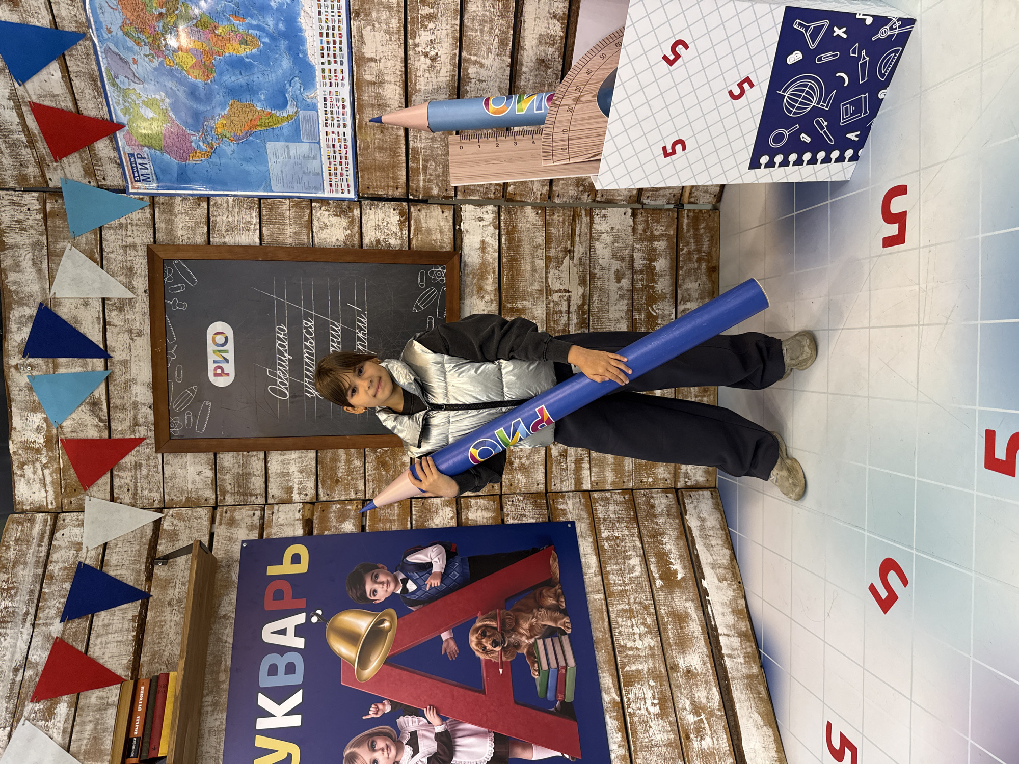 A young person stands in a studio decorated to resemble a classroom. The person is holding a large blue pencil prop and is wearing a silver puffer vest over a dark shirt and dark pants. The background features a wooden wall with a bookshelf containing several books on the left, a large poster with the Cyrillic word "БУКВАРЬ" (Bukvar, meaning alphabet book) and animated children, and a chalkboard with the word "РИО" (RIO) and a handwritten message in Russian. To the right of the chalkboard is a world map. In front of the person, on the floor, are red numbers "5" painted on a grid pattern. A large, box-like prop resembling a notebook with school-related drawings is also present. The overall scene suggests a themed event or set related to education or back-to-school activities. The location is identified as Korobovo, Russia.