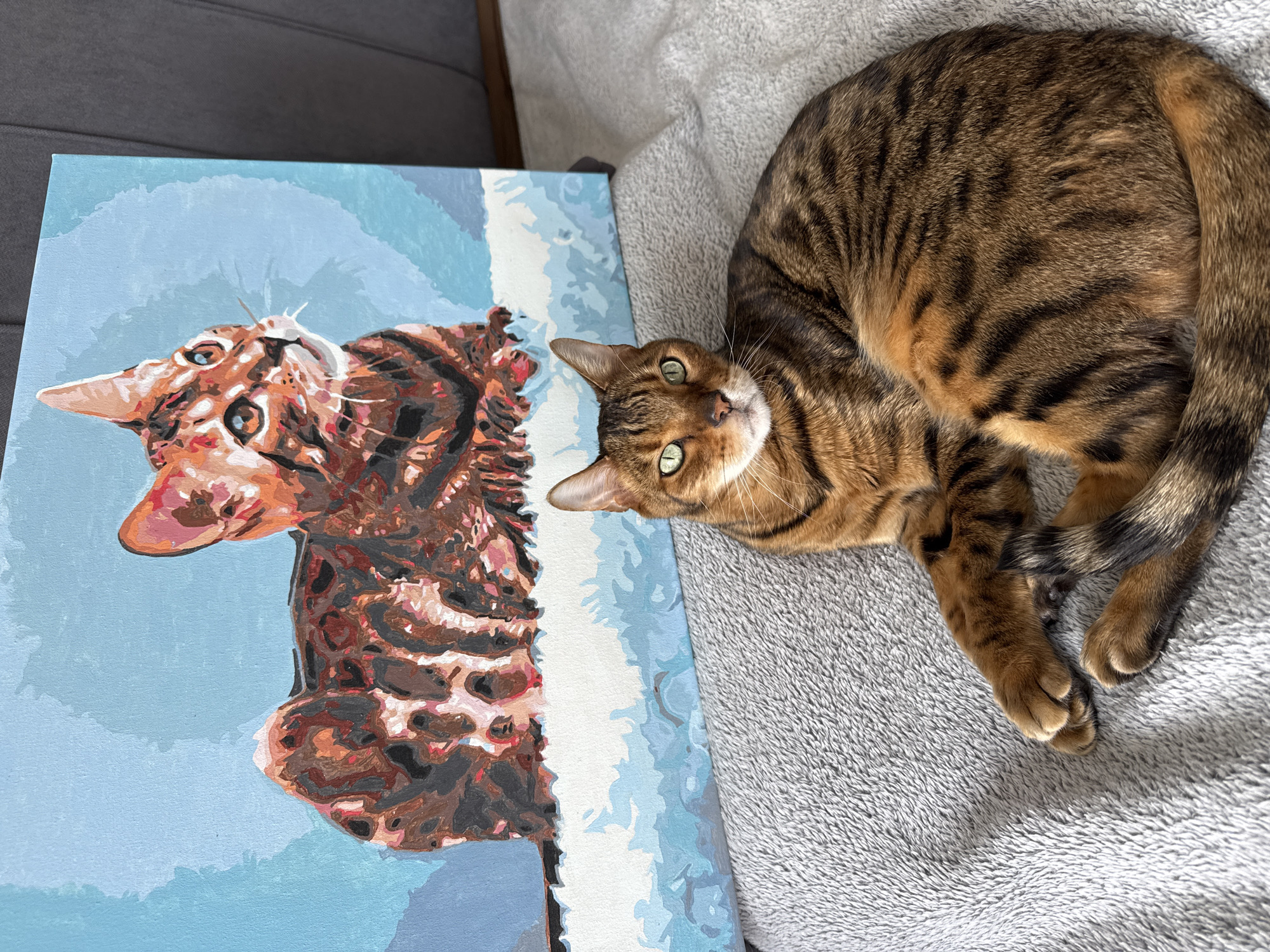 An overhead shot captures a Bengal cat resting on a grey, plush blanket next to a stylized painting of a similar cat. The real cat, positioned on the right side of the frame, is curled up with its head turned, looking directly at the viewer with bright green eyes. Its fur displays distinct brown and black rosetted patterns, characteristic of the breed.
To the left of the live cat, a canvas painting leans against an unseen surface. The painting features a cat with strikingly similar markings, rendered in a blocky, paint-by-numbers aesthetic. The painted cat is oriented vertically, seemingly looking upwards, and is set against a light blue background with abstract white and darker blue shapes, possibly depicting water or clouds. The artwork captures the cat's prominent green eyes and ear shape.
The scene appears to be indoors, likely in a comfortable home setting, with soft, diffused lighting illuminating both the cat and the artwork. The overall impression is one of a beloved pet being immortalized in art, with the live subject posing casually beside its painted likeness. No visible text is present in the image.