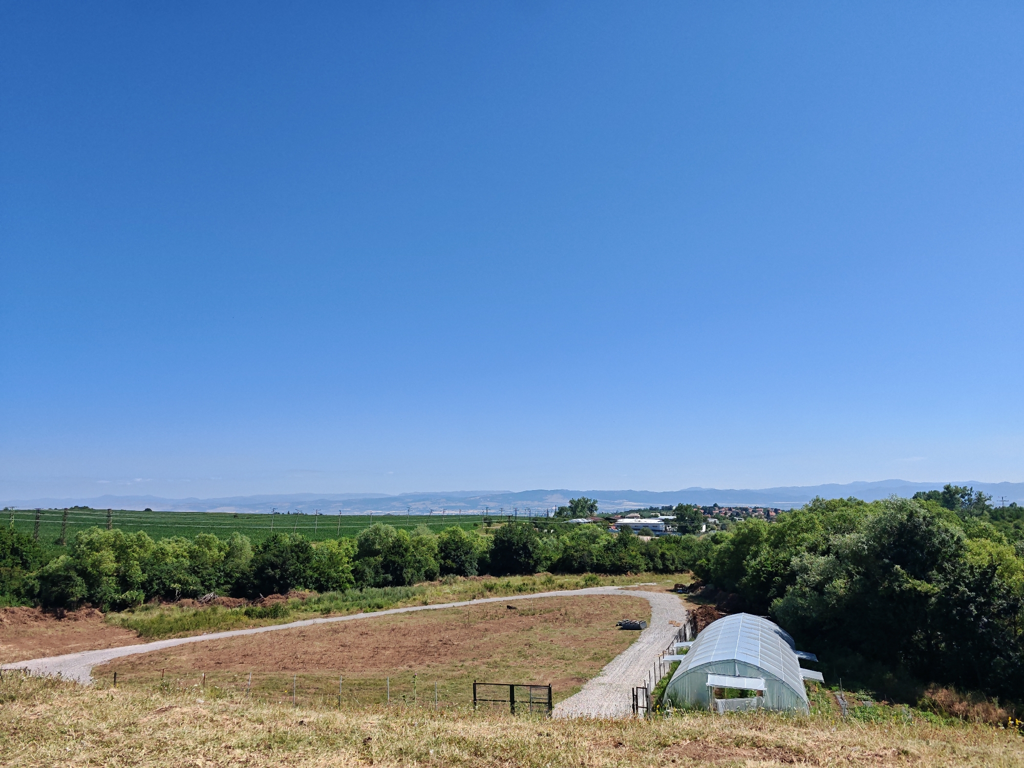 The image depicts an outdoor scene with a vast, clear blue sky dominating the upper half. In the foreground, a dry, grassy slope transitions into a dirt path curving through a brown, fallow field. A small, dark metal fence is visible in the field. To the right, a modern, arched greenhouse with a translucent, curved roof sits next to the dirt path. A gravel path leads to the greenhouse, and some construction materials are piled nearby. Beyond the field and greenhouse, a line of lush green trees forms a dense barrier. Further in the background, more trees and vegetation are visible, interspersed with agricultural fields and distant buildings that suggest a town or village. Power lines and transmission towers dot the landscape in the middle ground, indicating infrastructure. The horizon is lined with hazy, undulating hills or mountains. The scene appears to be set in a rural or semi-rural area, possibly on the outskirts of Ivanyane, Bulgaria, given the context provided. The overall impression is one of open space and agricultural activity under a bright, sunny sky.