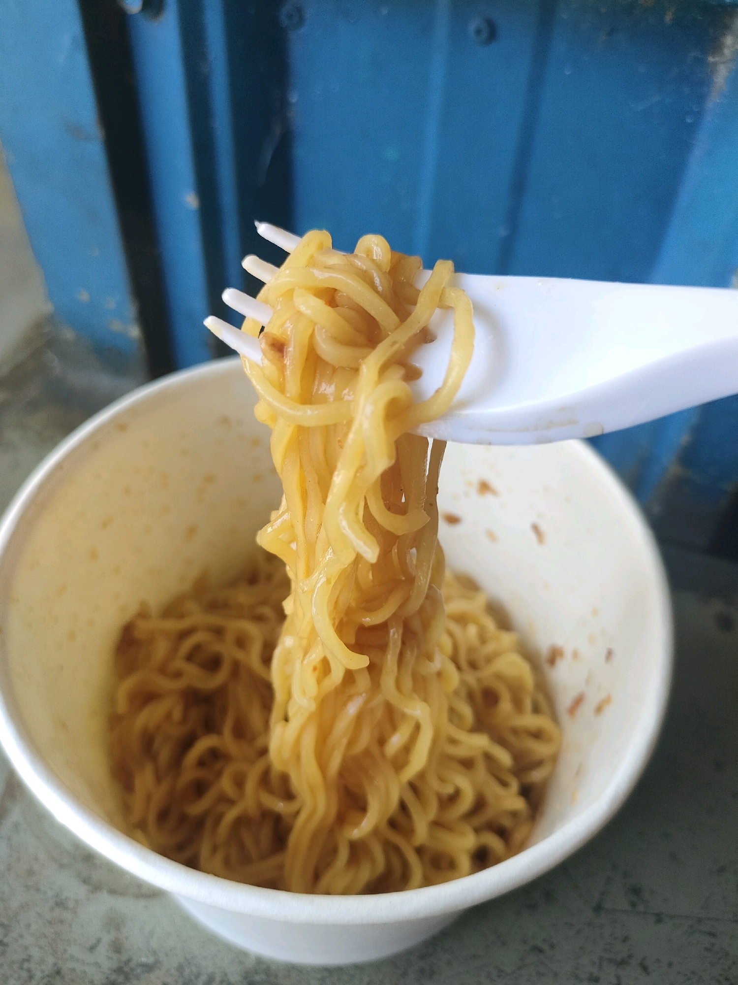 A white plastic fork is lifting a clump of cooked instant noodles from a white paper cup. The noodles are golden yellow and appear slightly oily. The cup is partially filled with more noodles. The background is out of focus, but shows vertical blue panels, suggesting an outdoor or industrial setting. The surface beneath the cup is a mottled grey. The image was taken in Sukodono, Indonesia.