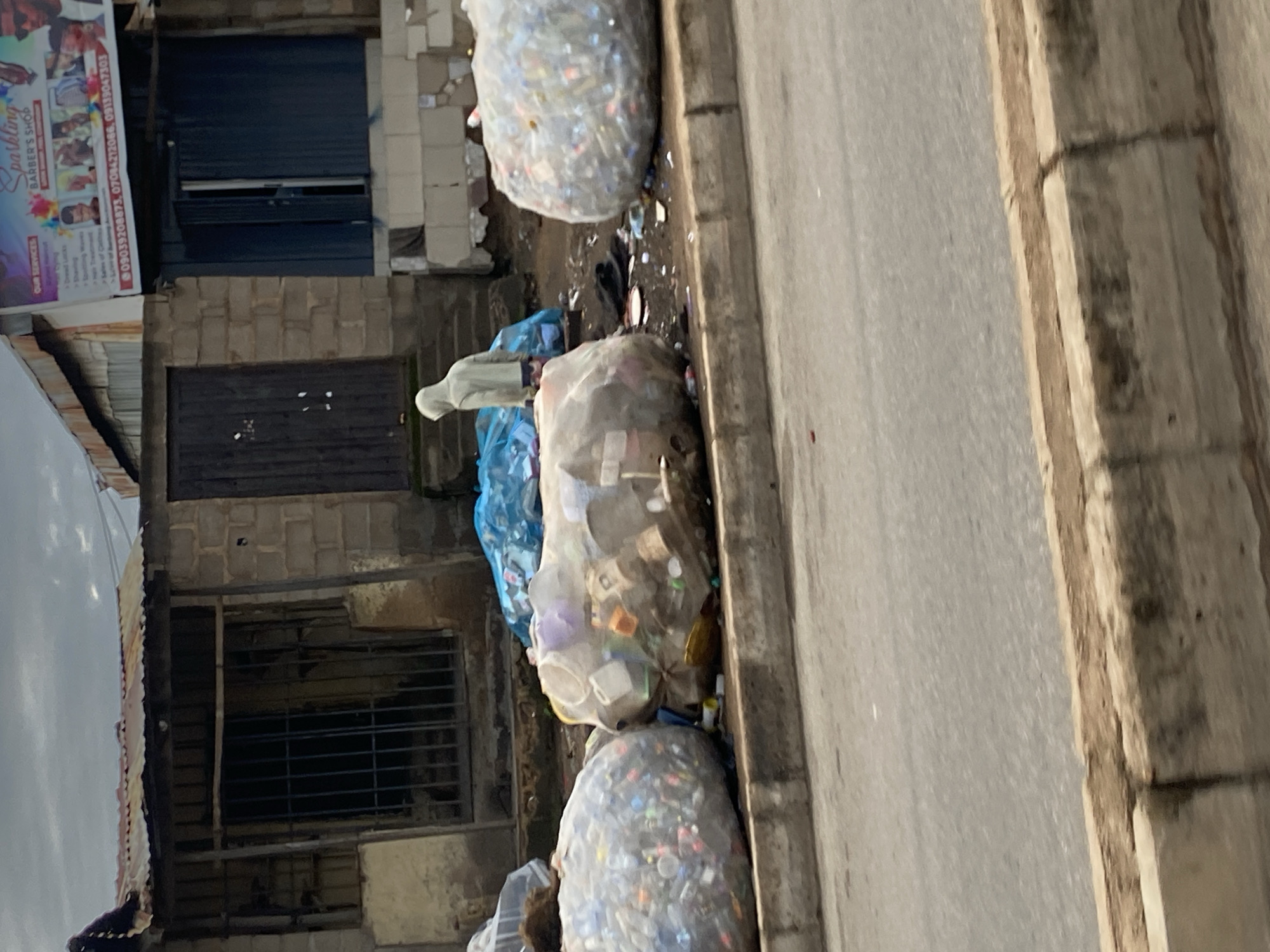 In the city of Barakin Lerawi, Nigeria, a person wearing a grey hooded sweatshirt stands in front of a collection of large, clear plastic bags filled with discarded plastic bottles and other refuse. The bags are piled on the side of a paved road, near a weathered building with a corrugated metal roof and brick walls. A blue tarp covers a pile of waste to the left of the person. A sign advertising "Sparkling Barber's Shop" is visible on the building's facade, along with contact information. The ground is littered with more debris, and the sky is overcast.