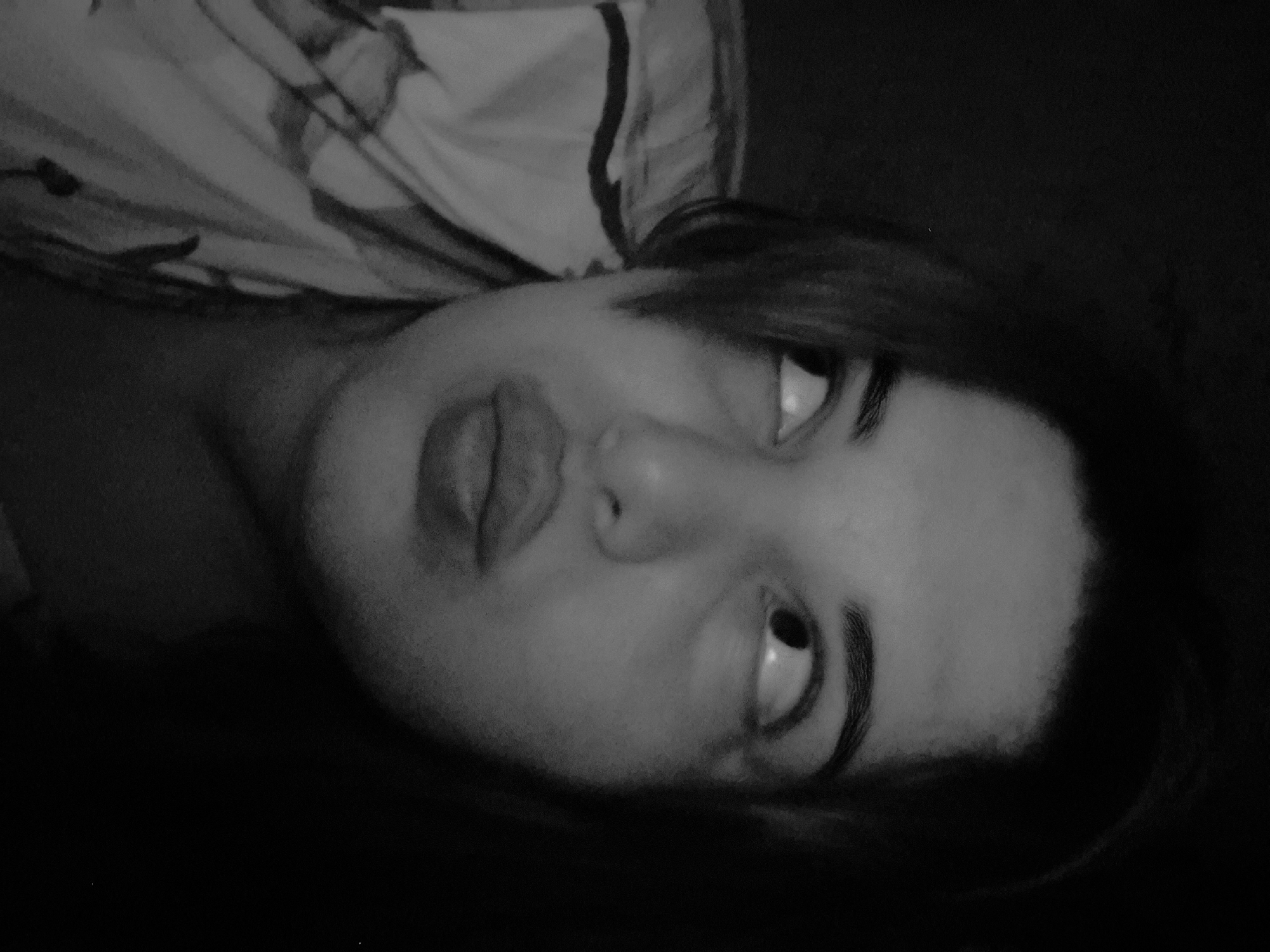 A close-up, black-and-white photograph shows a person's face, possibly female, tilted at an angle. The subject is looking upwards with both eyes. Their lips are slightly puckered. A patterned fabric, possibly clothing or bedding, is visible in the background behind their head. The rest of the background is dark and indistinct. There is no evidence of a location in the city of Амзя, Russia.