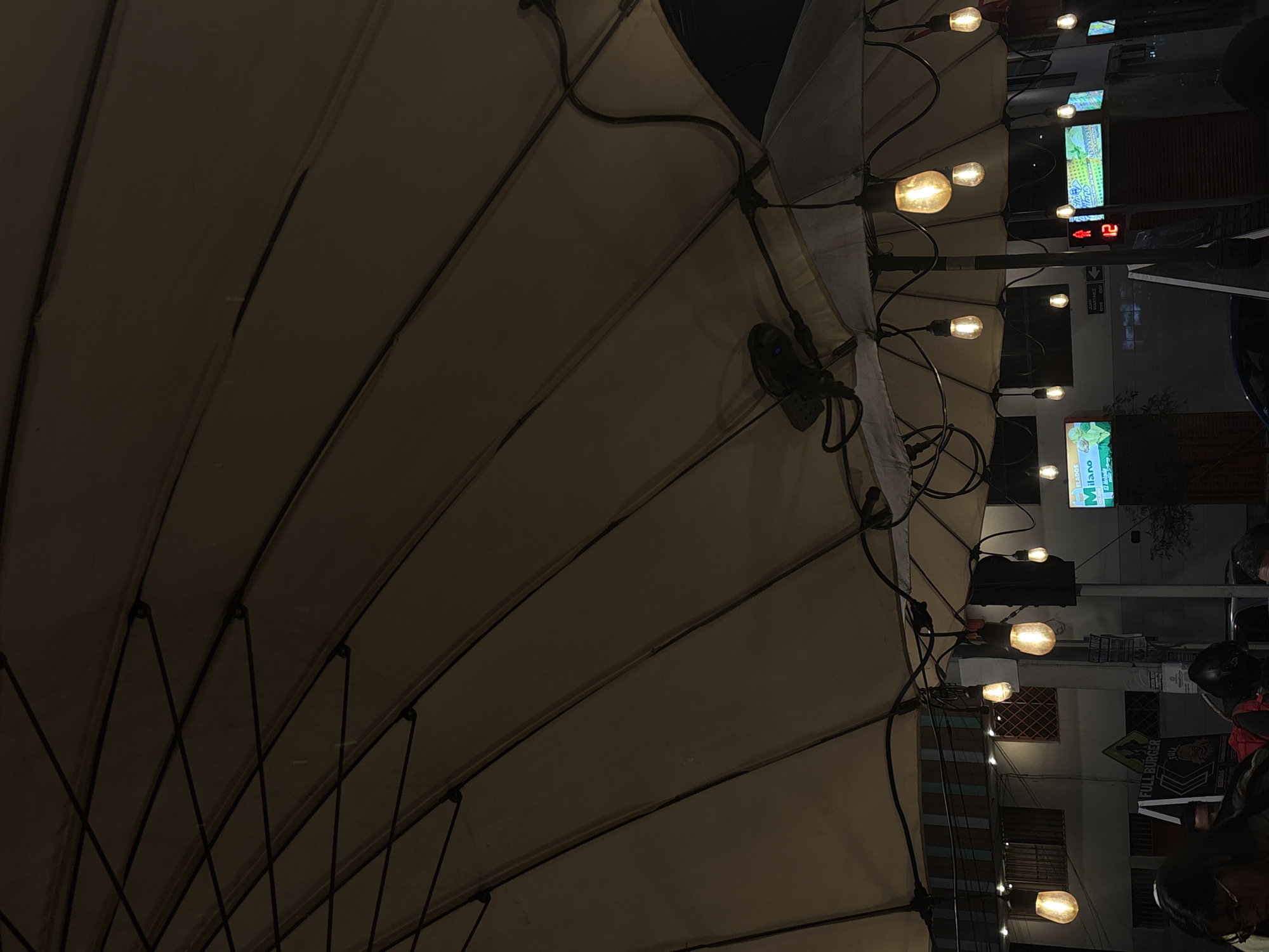 The image captures an outdoor seating area at night, likely a restaurant or cafe, beneath a large, taut, light-colored umbrella or awning. The fabric of the umbrella is supported by a metal frame, with visible ribs and crossbeams. A string of exposed Edison-style light bulbs illuminates the scene, casting a warm glow. Some of these bulbs are partially obscured by the umbrella's structure. A small black device, possibly a fan or security camera, is mounted on the underside of the umbrella.
In the background, buildings are visible, some with windows. Signage is present, including a green sign with "MILANO" and images of ice cream, and another sign with "FULL BURGER." A traffic light shows a red pedestrian signal and the number "12" illuminated. The street appears to be paved.
The lower portion of the image shows the heads and shoulders of several people, implying a social gathering. One individual wears glasses. The atmosphere suggests a cool, possibly damp evening, as indicated by the subdued lighting and the slightly misty appearance in the background. The specific location in Iquique, Chile, is not explicitly shown but the presence of the signs and general urban setting are consistent with a city environment.