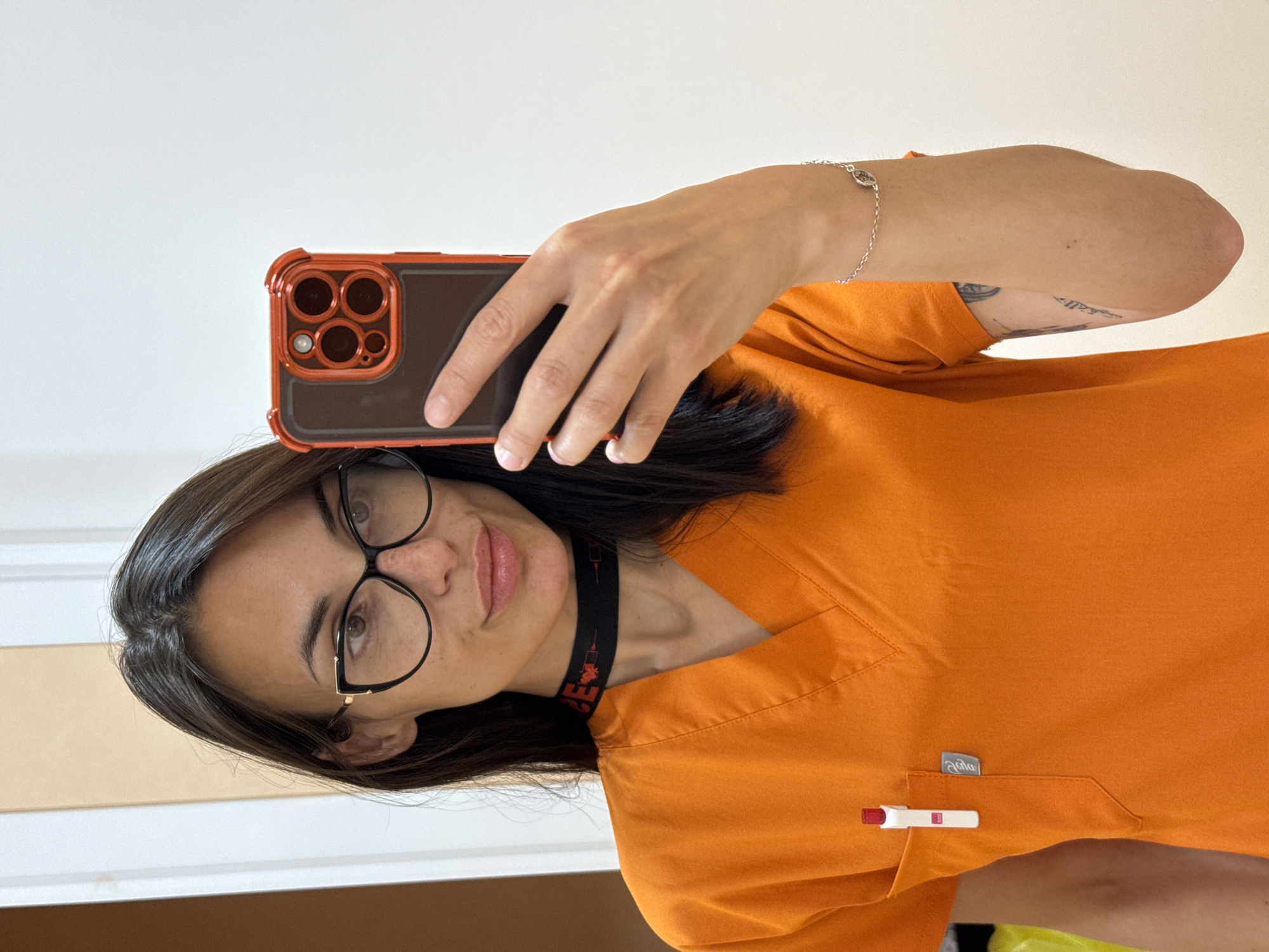 A woman is taking a selfie indoors. She is wearing black-rimmed glasses, a black choker with a medical symbol design, and an orange scrub top. She has a silver bracelet on her wrist. A pen is clipped to the scrub top pocket. She is holding a smartphone with a red-trimmed case. The background is out of focus and shows a white wall. The image orientation is rotated 90 degrees clockwise. There is no indication of the location being in the city of Pleven, Bulgaria.