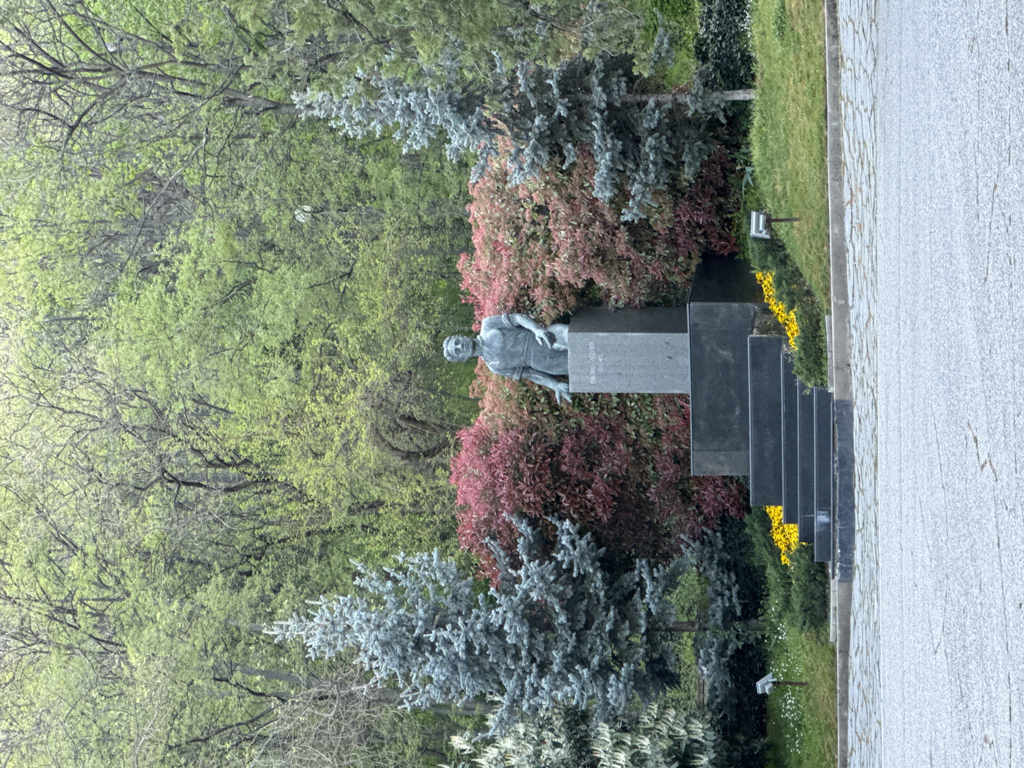 The image shows the Monument of the Soviet Army in Plovdiv, Bulgaria. A bronze sculpture of a soldier in work clothes sits atop a gray stone plinth, which is set in front of a variety of shrubbery with red, green, and blue-gray foliage. There are five additional smaller rectangular monuments lined behind the statue on the left. Yellow flowers grow near the base of the monument along the side of a paved pathway. In the background are trees with green foliage.