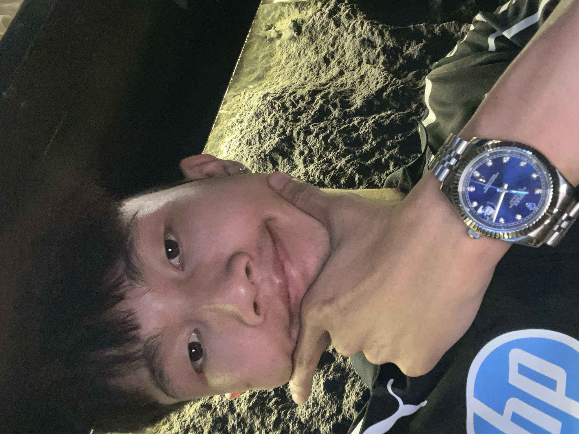 An overhead shot captures a person, likely male, tilted in the frame, looking towards the camera. He wears a black t-shirt featuring a prominent white "HP" logo and white stripes on the sleeve. A silver-toned wristwatch, appearing to be a Rolex Datejust with a blue dial, diamond hour markers, and a jubilee bracelet, adorns his left wrist. The watch dial clearly shows "ROLEX OYSTER PERPETUAL DATEJUST SUPERLATIVE CHRONOMETER OFFICIALLY CERTIFIED" and "SWISS MADE" along with the Rolex crown.
The person's left hand is positioned under his chin and jawline, gently squeezing his cheek, creating a slightly playful expression with a subtle smile. His dark hair is visible, framing his face.
The background consists of a dark, highly textured surface, resembling a rough rock wall or cavern, illuminated from the left by a warm, yellow light source. This artificial lighting creates strong shadows and highlights on the rugged texture. The overall setting appears dark, suggesting an indoor environment or nighttime, possibly a themed space or a natural cave. The top left portion of the image is completely dark, suggesting an enclosure. Given the location context of Đà Nẵng, Vietnam, this could be a grotto, cave, or an elaborately designed interior.