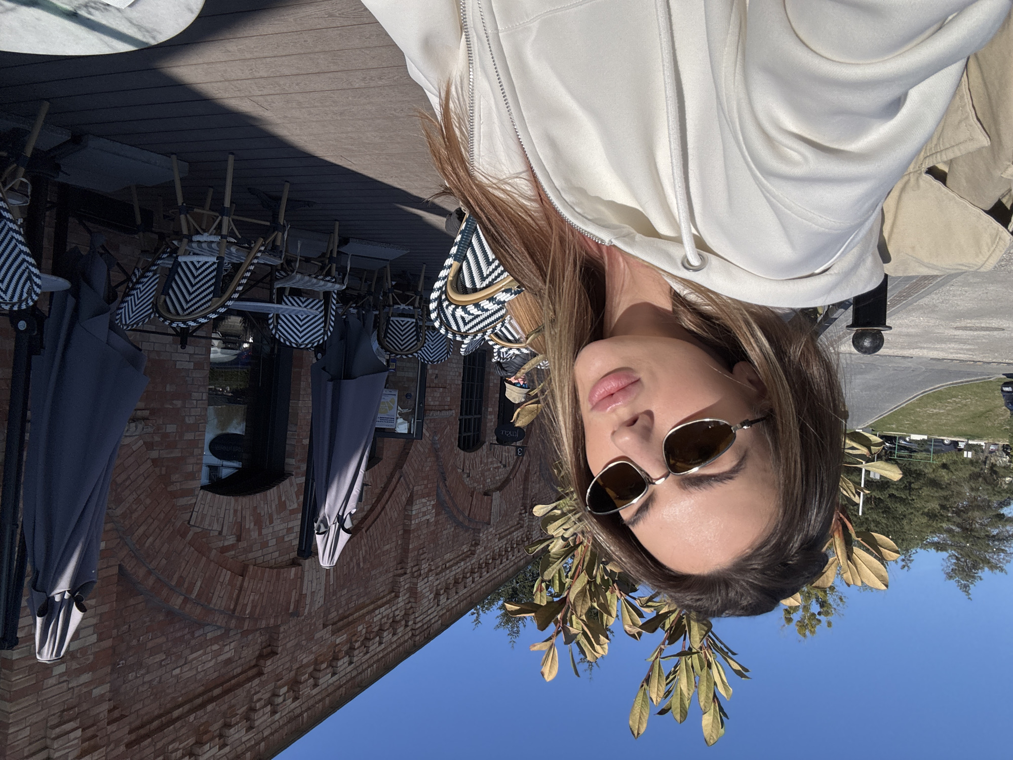 This image captures a young woman in an outdoor setting, likely a cafe or restaurant patio in Warsaw, Poland. The photograph appears to be a selfie or a close-up shot, with the woman looking directly at the camera.
**Subjects:** The primary subject is a young woman, visible from the chest up, positioned on the right side of the frame. She has light brown hair, is wearing stylish silver-framed sunglasses that obscure her eyes, and a light-colored (off-white or cream) zip-up hoodie. Her lips are slightly parted, with a glossy finish. Behind her shoulder, a patterned bag with a black and white chevron or zig-zag design is partially visible. Framing her face on the left is a branch with greenish-yellow leaves.
**Scene and Setting:** The background features an outdoor patio with a wooden deck floor. Numerous outdoor cafe chairs, characterized by black and white woven patterns on their backs and seats and light-colored frames, are visible; many appear stacked or arranged for storage. Several large, dark-grey closed patio umbrellas are also present. Behind the patio is a building constructed from red bricks, exhibiting classic brickwork and arched window details. The sky above is a clear, bright blue, with some distant trees and greenery on the horizon. A hint of what might be a road or path is discernible in the far background.
**Activity or Event:** The image captures a casual moment outdoors, with the woman posing for the camera.
**Notable Details:** The bright, direct sunlight suggests it is daytime, likely late morning or afternoon, casting clear shadows on the wooden deck. The weather appears sunny and pleasant. The combination of the cafe setting, brick architecture, and clear sky creates a leisurely urban ambiance. While the woman's eyes are hidden, her posture and slight smile suggest a relaxed and confident mood.
**Visible Text:** On a window or sign on the brick building, faint and partially obscured text is visible, possibly reading "now" or "noul," but it is too indistinct to be read clearly.