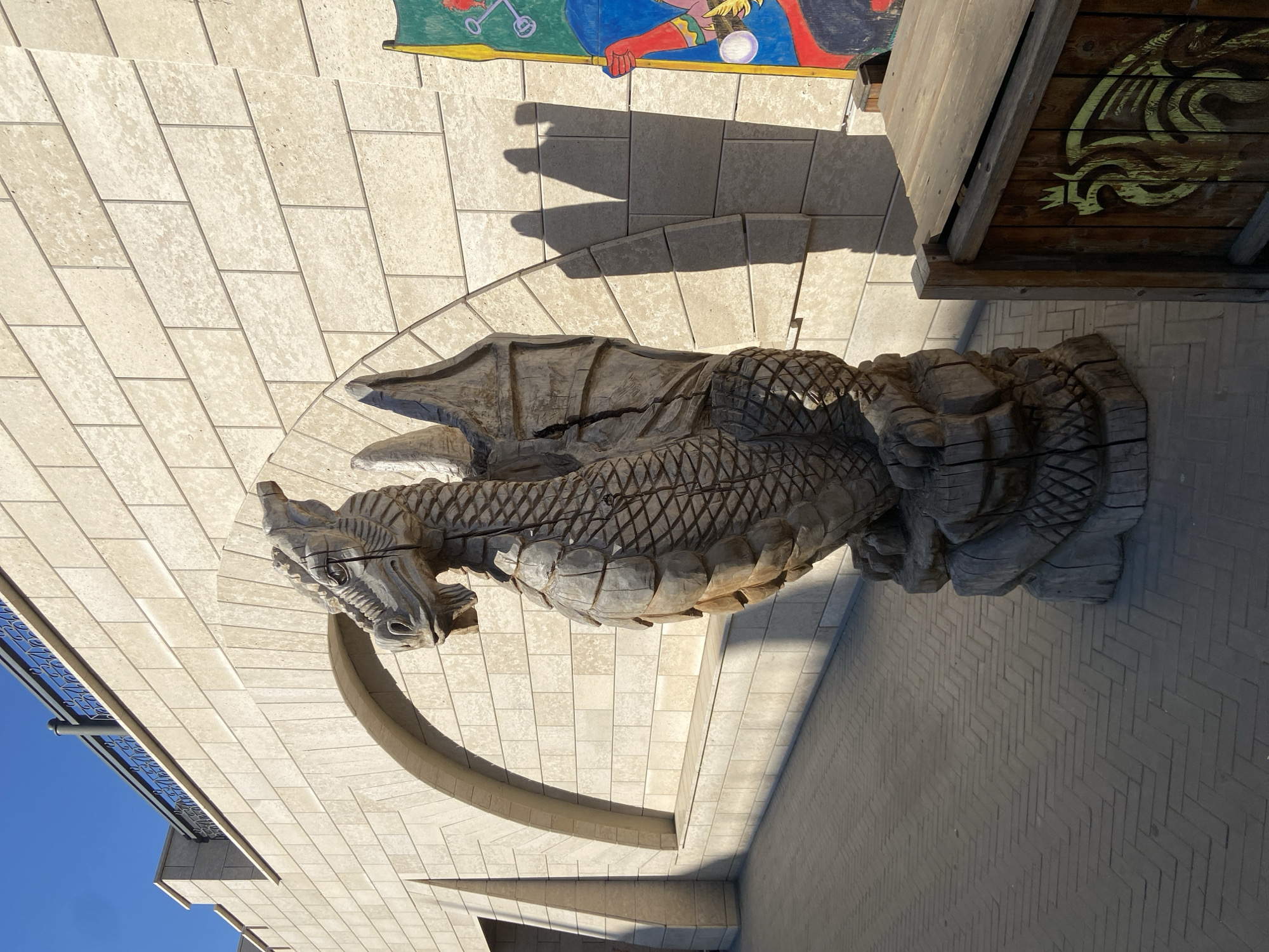A carved wooden dragon sculpture is the focal point, seemingly placed in a courtyard setting in Kazan, Russia. The dragon is depicted in detail, with scales and wings clearly visible. The courtyard is paved with light-colored stone or concrete blocks laid in a rectangular pattern. An arched architectural element is visible near the dragon's head. Other elements in the scene include a building with a blue railing, a shadow cast by the dragon, a wooden structure with a dragon motif, and a colorful mural in the background.