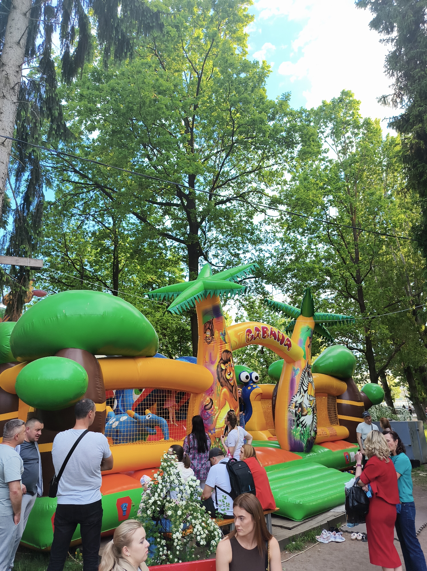 A daytime shot depicts an outdoor scene in Ternopil, Ukraine, featuring a large, inflatable "CABANA" jungle-themed bouncy castle surrounded by a group of people. The bouncy castle is predominantly green, orange, and yellow, designed with depictions of jungle animals like lions, zebras, and cartoonish blue elephants. Several adults stand around the inflatable structure, some seemingly watching children inside. A woman in a red dress is on the right side of the scene. There's a bench in the foreground where people are sitting. A planter filled with white flowers is positioned near the front of the bouncy castle. Tall green trees surround the area, providing a backdrop under a partly cloudy sky. The setting appears to be a park or recreational area within the city.