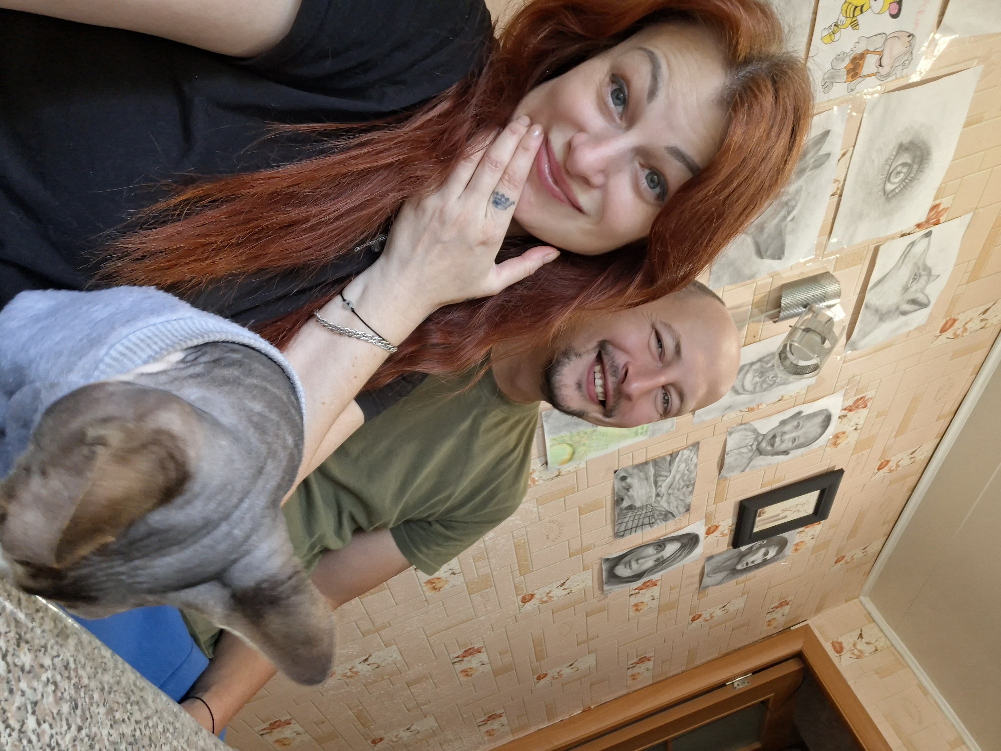 In Myadzyel, Belarus, a woman with vibrant red hair and a man with a shaved head are posing for a selfie in front of a wall adorned with various pencil sketches. The woman is in the foreground, her left hand resting on her chin, revealing a small crown tattoo on her ring finger and a silver bracelet on her wrist. She is wearing a black t-shirt. The man, in a green t-shirt, is smiling broadly beside her. Between them and partially obscuring the lower half of their bodies is a Sphynx cat wearing a light blue sweater. The wall behind them features several framed and unframed drawings, including a detailed eye, a wolf, a tiger, a prehistoric human, two portraits of children, a landscape, and a portrait of a woman. A metallic object, possibly a lamp, is mounted on the wall. The overall environment suggests a home or studio setting with a warm, personal touch due to the artwork.
