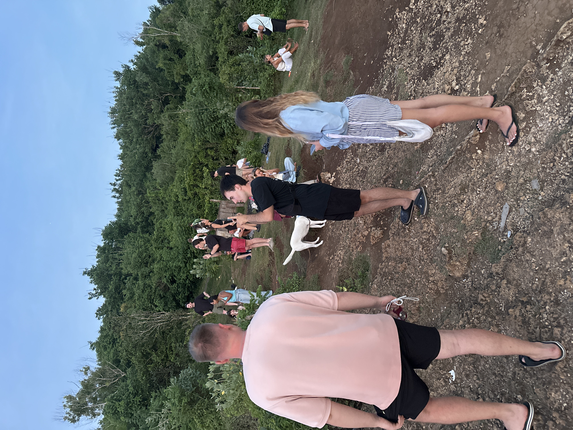 The scene shows a group of people gathered on a rocky, uneven terrain with a dense green forest as a backdrop. The sky is a pale blue. Several individuals are looking at their phones, suggesting a casual gathering or a planned event. Two white dogs are present among the group. The location appears to be outdoors in Kuta Selatan, Indonesia, given the vegetation and rocky terrain. The clothing worn by the individuals is casual, indicating a relaxed atmosphere. The lighting suggests that it might be late afternoon or early evening.