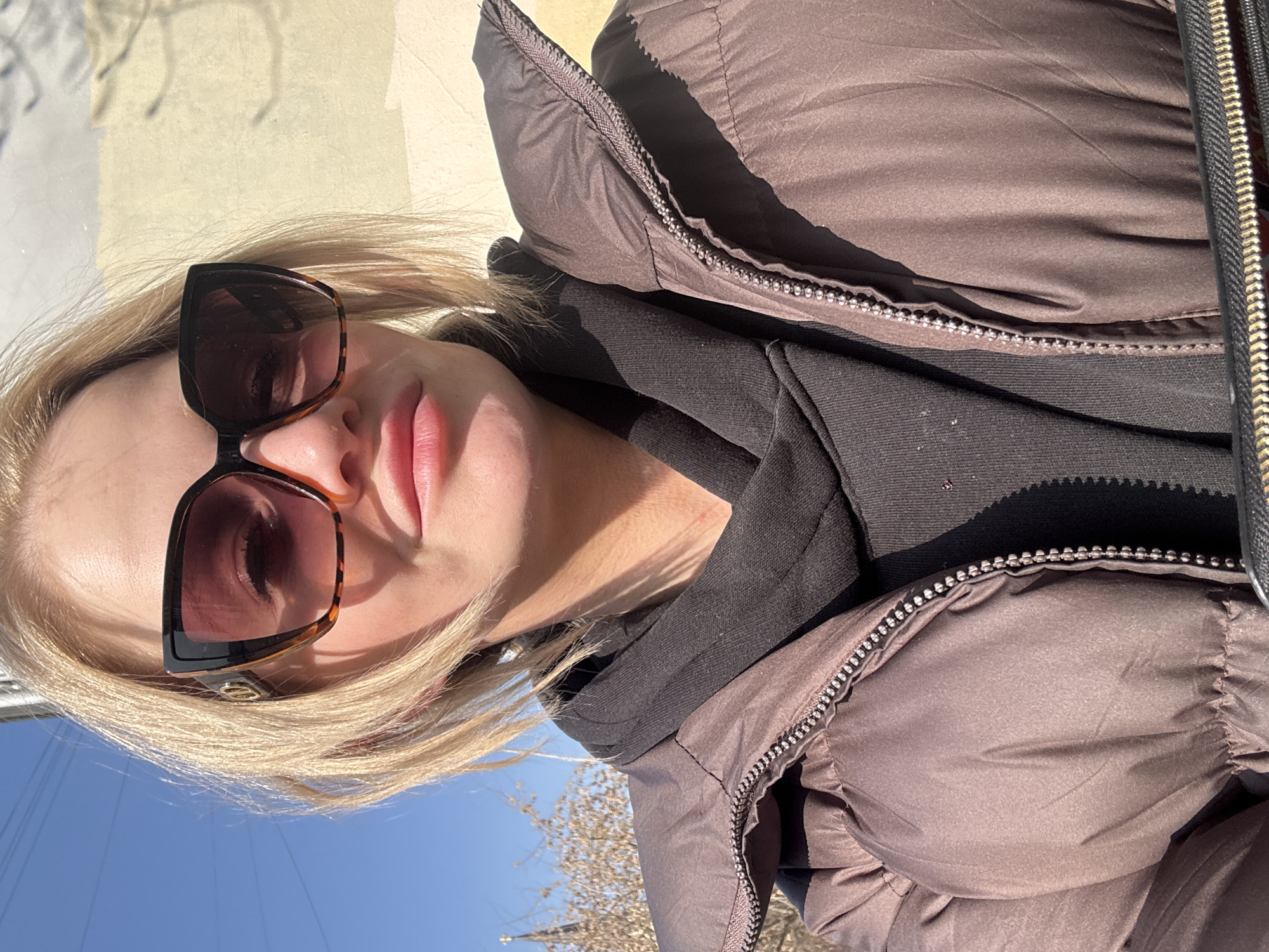 A woman wearing sunglasses and a brown puffer jacket with a black hoodie underneath takes a selfie outdoors. The background suggests a sunny day. The location is likely Saint Petersburg, Russia based on the architectural style in the upper left corner of the picture.