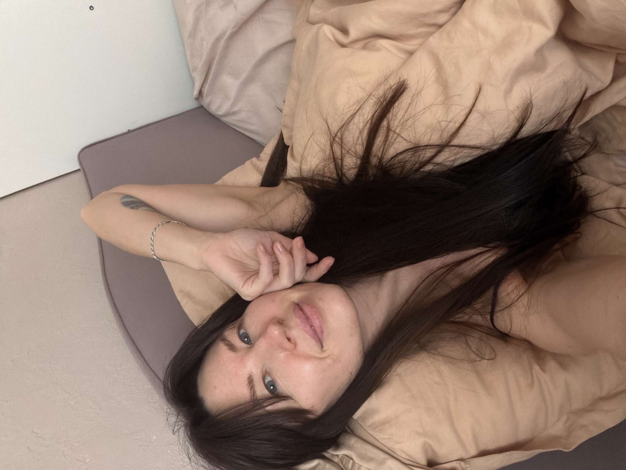 A person lies on a bed in a room with cream-colored bedding. The individual has long, dark hair spread across the pillow, pale skin, and blue eyes. They are wearing a thin silver bracelet on their left wrist. A tattoo is visible on their left arm. The bed has a grey headboard against a white wall. The location is implied to be in Perm, Russia based on the user's request, but no location-specific details are discernible in the image itself.