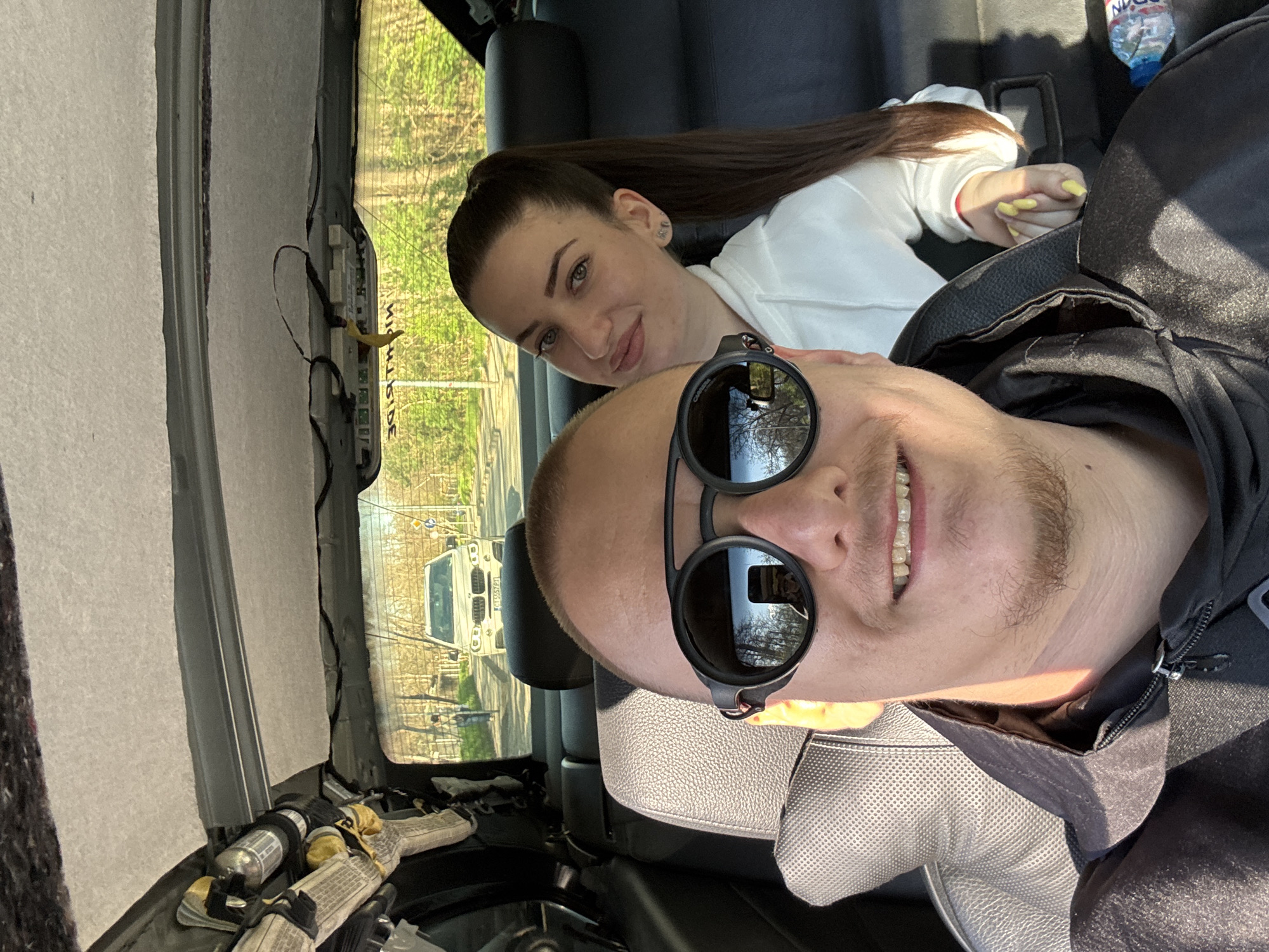 The image captures a selfie inside a car, likely during or after car repair. A man in sunglasses, wearing a black jacket, is at the forefront, smiling. Behind him lies a woman with her hair in a ponytail, wearing a white top, looking at the camera. The interior of the car shows exposed parts, potentially related to repairs. Through the rear window, a street scene is visible, featuring a light-colored car and trees, suggesting an outdoor environment. The license plate visible in the reflection is from Plovdiv, Bulgaria. There is also a water bottle near the top of the frame.