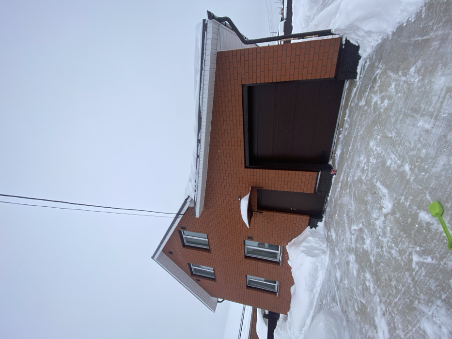 This image captures the exterior of a two-story residential house with an attached garage in Kultaevo, Russia, under an overcast sky during winter. The building features reddish-brown brickwork with a dark brown base trim and a dark brown pitched roof, which is partially covered in white snow. White soffit and fascia are visible along the roofline, with a dark brown gutter and downspout on the right. Four rectangular windows with white frames are visible on the front facade: two on the upper story and two on the lower story. A dark brown pedestrian door, situated to the left of the garage, is sheltered by a small, rounded, snow-covered awning. The attached garage features a large, dark brown ribbed roll-up door. Overhead utility wires extend diagonally from the upper left towards the house. The foreground consists of a paved surface, partially cleared but still covered with a thin layer of snow and ice, leading up to the house and garage. Significant snowdrifts are accumulated against the foundation of the house and along the perimeter of the cleared area. A small, bright green plastic shovel or scoop lies on the partially cleared ground in the lower-left corner. A small red cylindrical object is visible at the base of the wall between the pedestrian door and the garage door. No individuals, actions, or direct interactions are depicted.