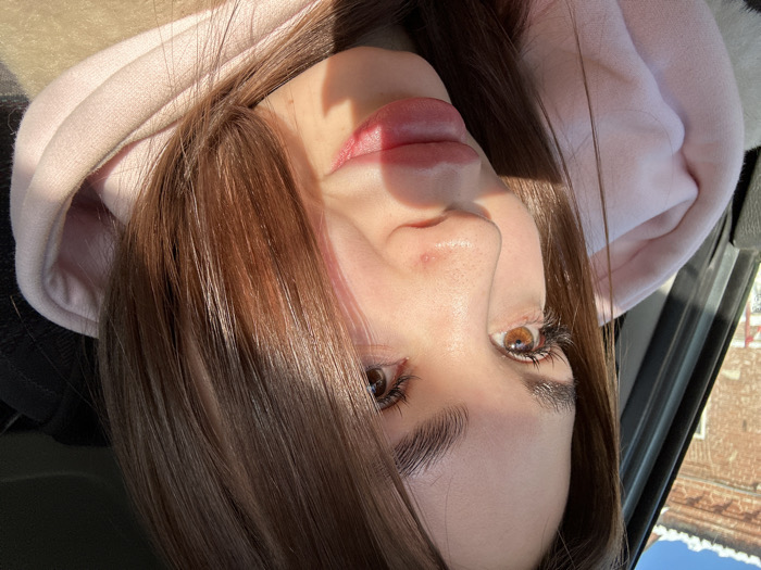 An extreme close-up, chest-up portrait captures a young adult female, likely positioned within a vehicle, in Votkinsk, Russia. The subject has long, straight brown hair, brown eyes with prominent eyelashes, and full, slightly parted, pinkish lips. Two small reddish blemishes are visible on the bridge of her nose. She is wearing a light pink hooded sweatshirt beneath a beige or light brown outer garment, visible on the right. Her head is angled slightly to the left, and she gazes upward and leftward. Direct sunlight brightly illuminates the left side of her face and hair, casting a distinct shadow across the right side of her face and neck. Through a window visible on the left, a blurred reddish-brown brick building and a clear blue sky are discernible in the background.
