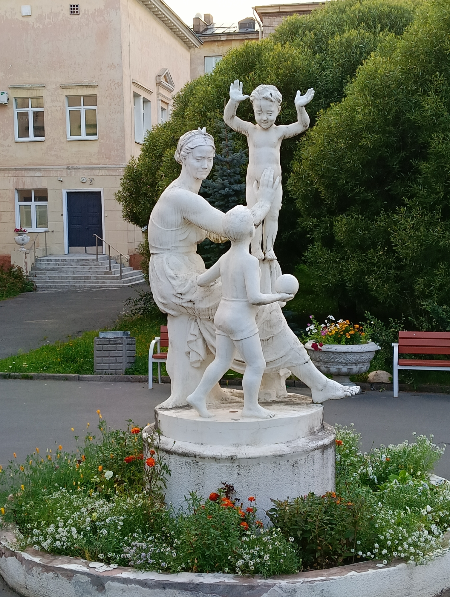 In Petrozavodsk, Russia, a white stucco statue depicts a woman with two children on a circular pedestal surrounded by a flower bed. The woman, seated, gazes at the children, one standing and one crouched, who appear to be playing. The standing child has arms raised, while the crouched child holds a ball. In the background, a building with a light-colored facade and white-trimmed windows, a dark blue door with a ramp leading to it, and lush green trees are visible. Two red-and-white park benches and a concrete planter with flowers are positioned to the right of the statue. The ground in front of the statue is paved, and a strip of grass with scattered wildflowers runs alongside it. A concrete trash receptacle is situated to the left of the statue.