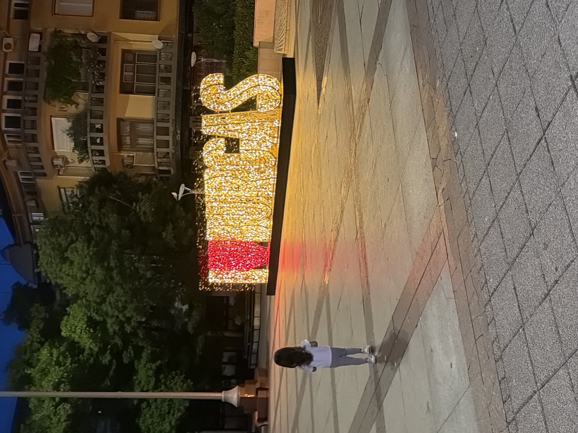 The image shows a child standing in front of a large, illuminated sign in Burgas, Bulgaria, which reads "I LOVE BURGAS". The sign is composed of large, stylized letters outlined with numerous bright lights, with the letter "U" being a vibrant red and the other letters a glowing yellow. The child, facing away from the camera, is wearing a white t-shirt and grey pants, standing on a paved plaza. In the background, a multi-story building with balconies is visible, and trees with dense foliage line the street. The scene is illuminated by the artificial lights of the sign and the ambient evening light, creating a warm and inviting atmosphere.